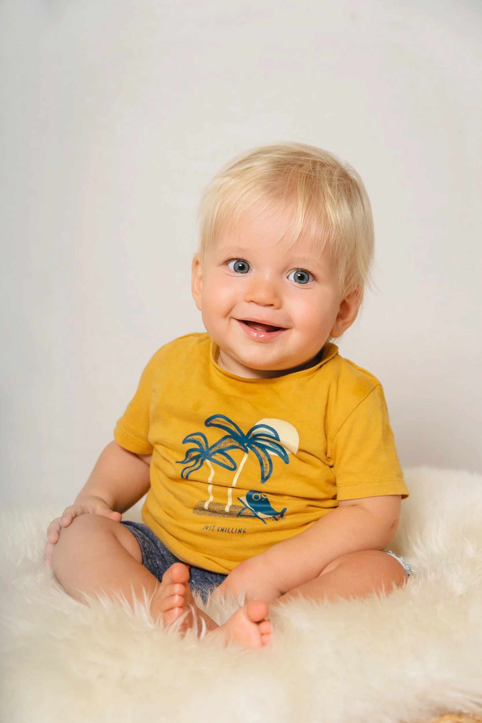 A young boy with blonde hair and blue eyes, smiling while sitting on a white fluffy surface, wearing a yellow T-shirt with a palm tree graphic and denim shorts.