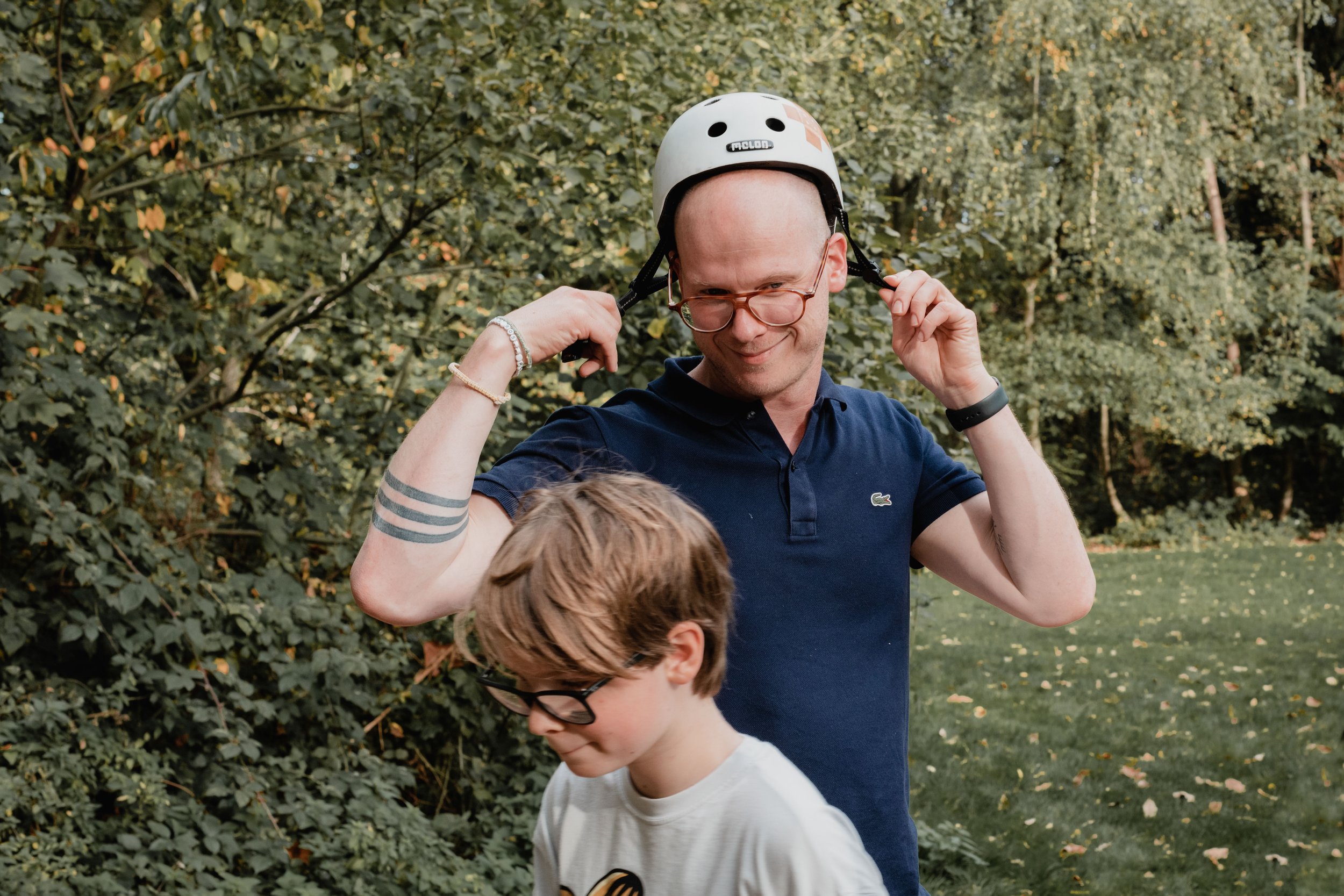 A man wearing glasses and a white helmet adjusts it as a child with glasses and a white shirt stands in front of him. They are outdoors near green bushes and trees.
