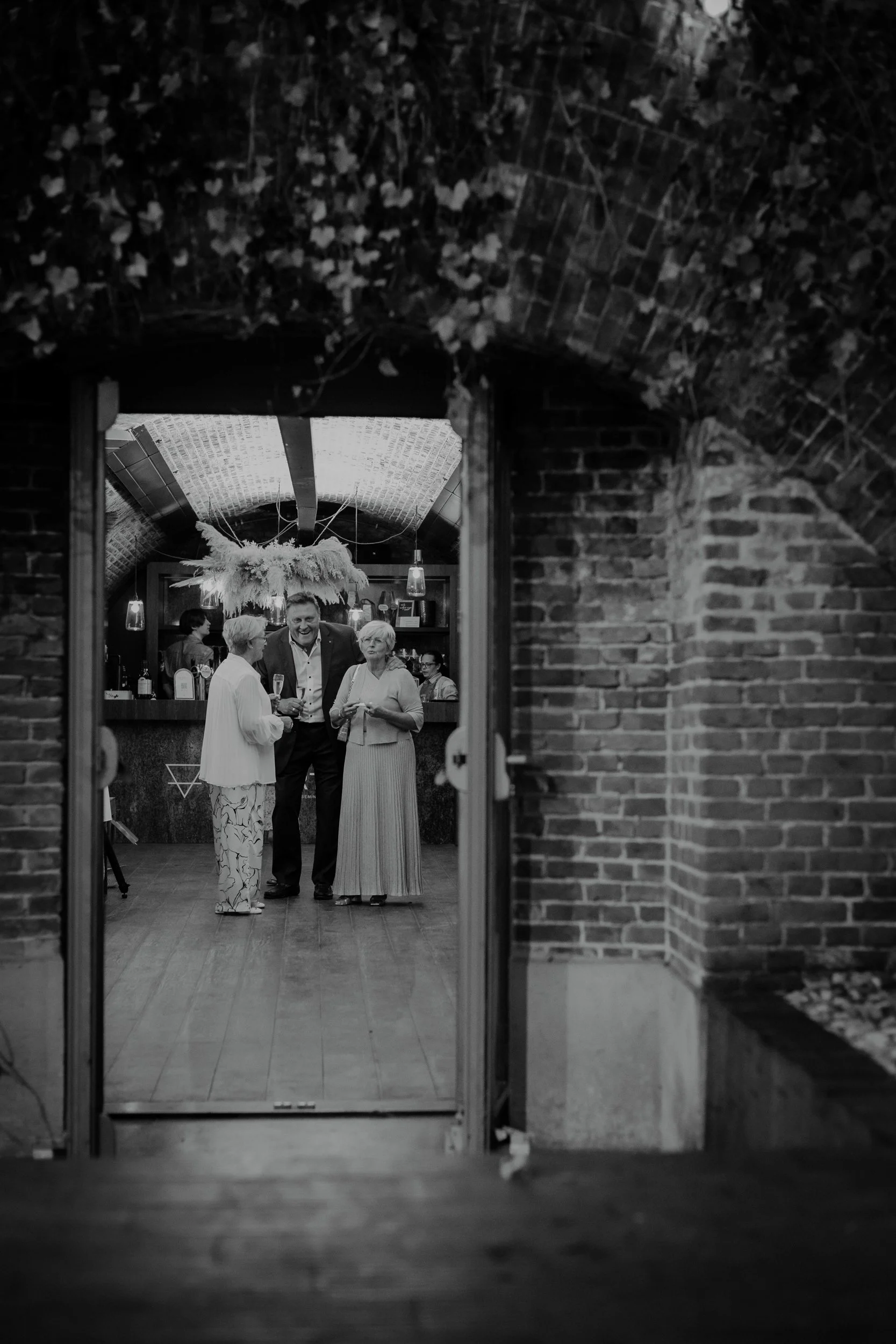 People gathered and talking at an indoor social event viewed through a doorframe, with brick walls and ceiling, and decorative hanging plants.