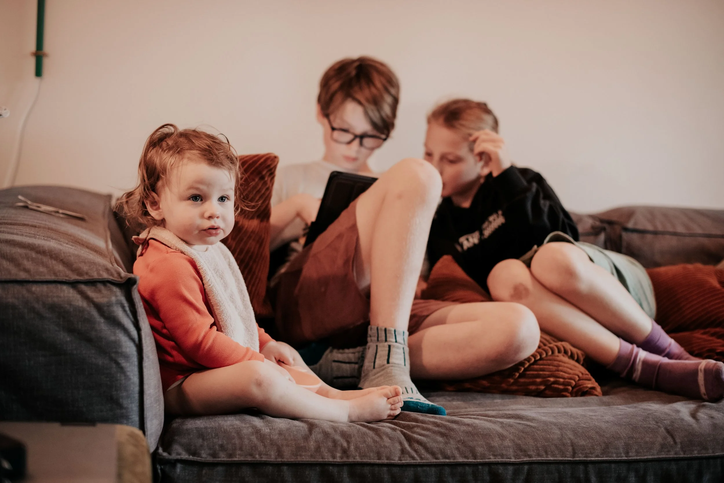 Three children sitting on a brown couch in a living room. The youngest, with curly hair, is in the foreground looking at the camera. The two older children are focused on a tablet and are sitting close together.