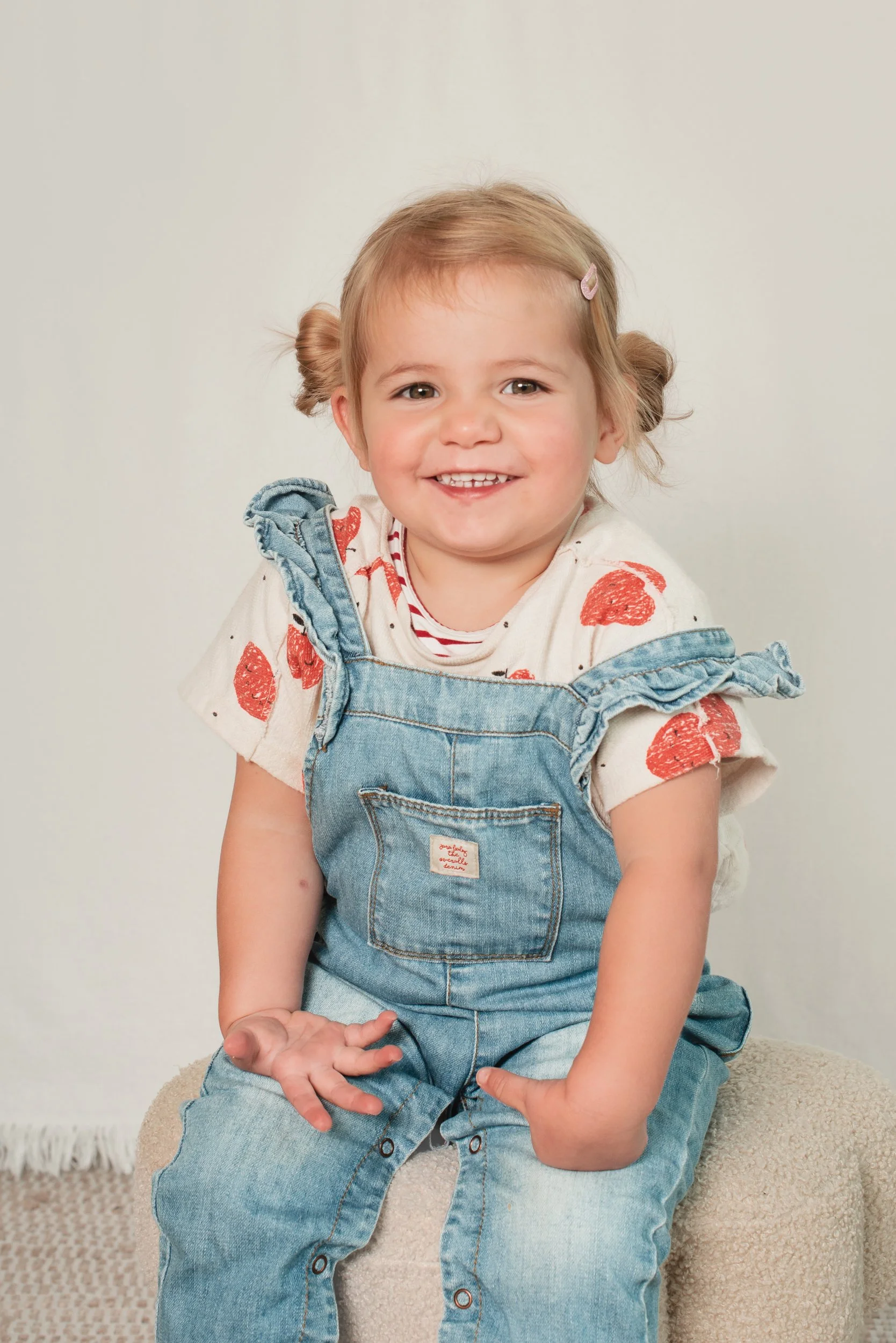 A smiling young girl with light brown hair in pigtails, wearing a white shirt with orange watermelon prints and denim overalls, sitting on a beige textured surface with a plain white wall background.