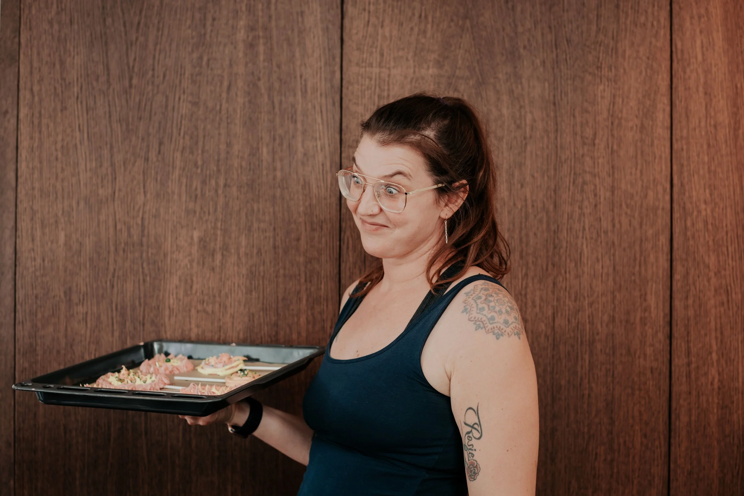 A woman with glasses and tattoos on her arm, wearing a sleeveless top, holding a tray of decorated cupcakes, standing against a wooden wall.