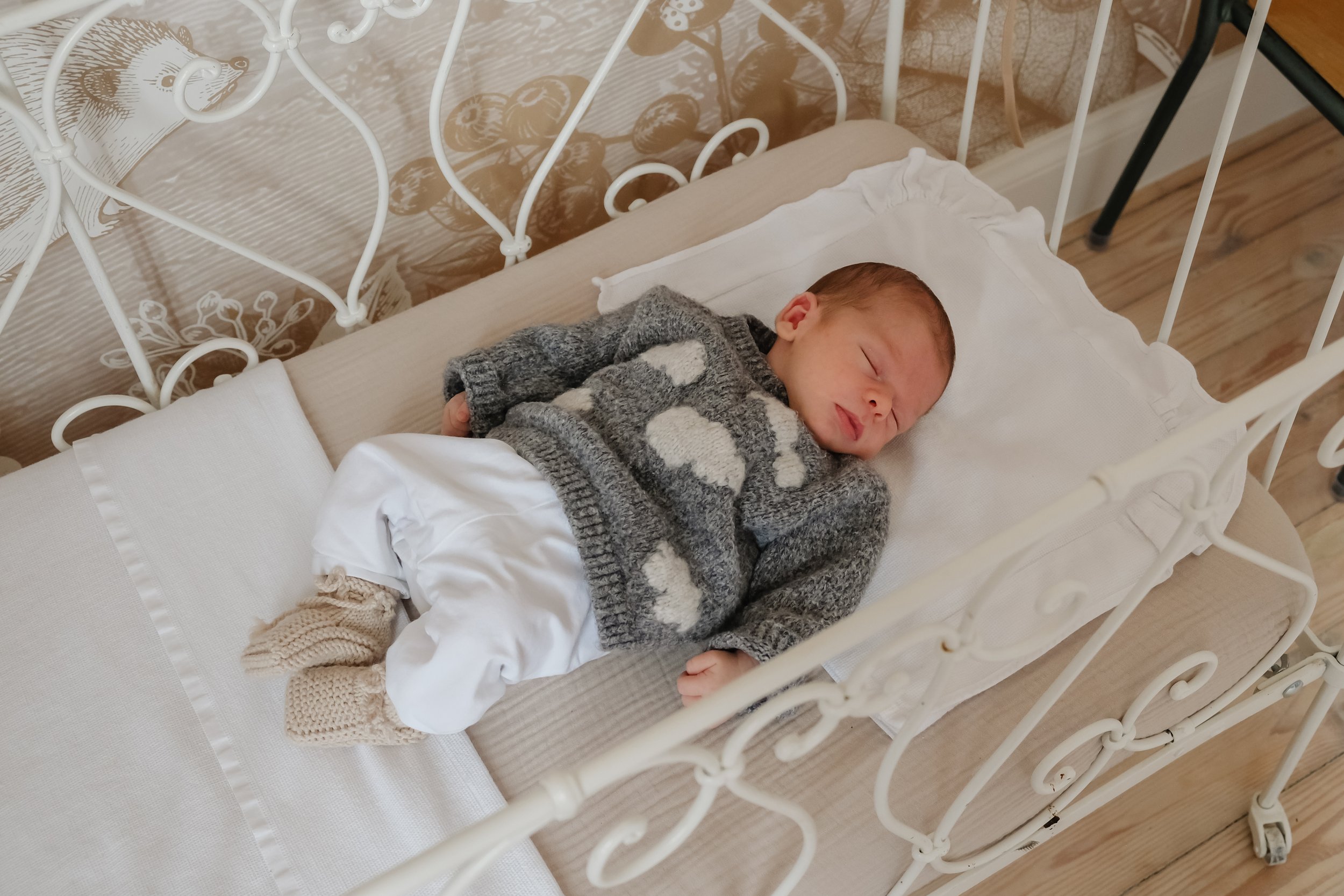 A sleeping infant with light skin and brown hair lying in a vintage white iron crib, dressed in a gray sweater with white polka dots and beige knitted socks, in a room with wooden flooring.
