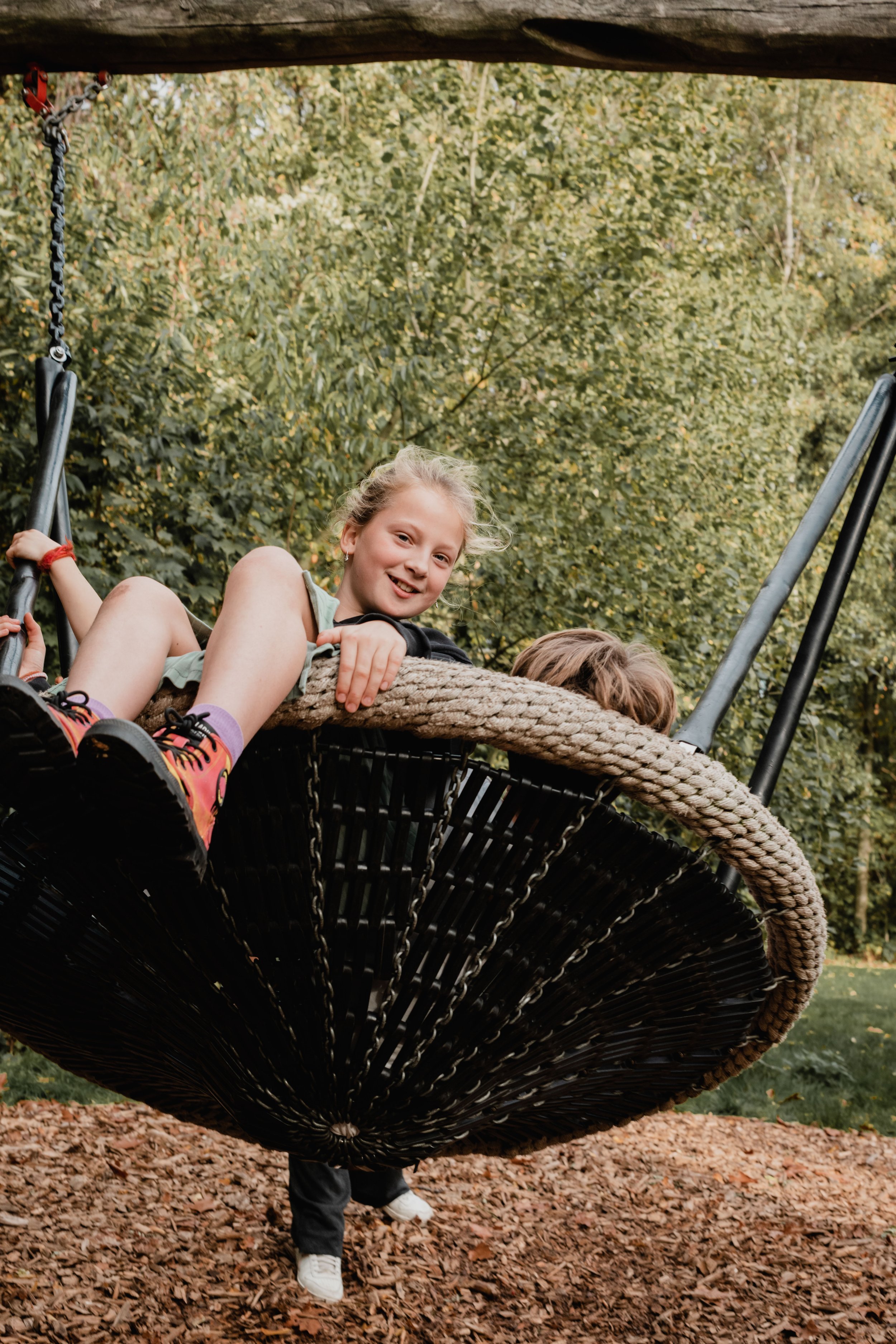 A young girl with blonde hair smiling and lying on a large rope swing basket outdoors, with her legs extended, surrounded by green trees and fallen leaves on the ground.