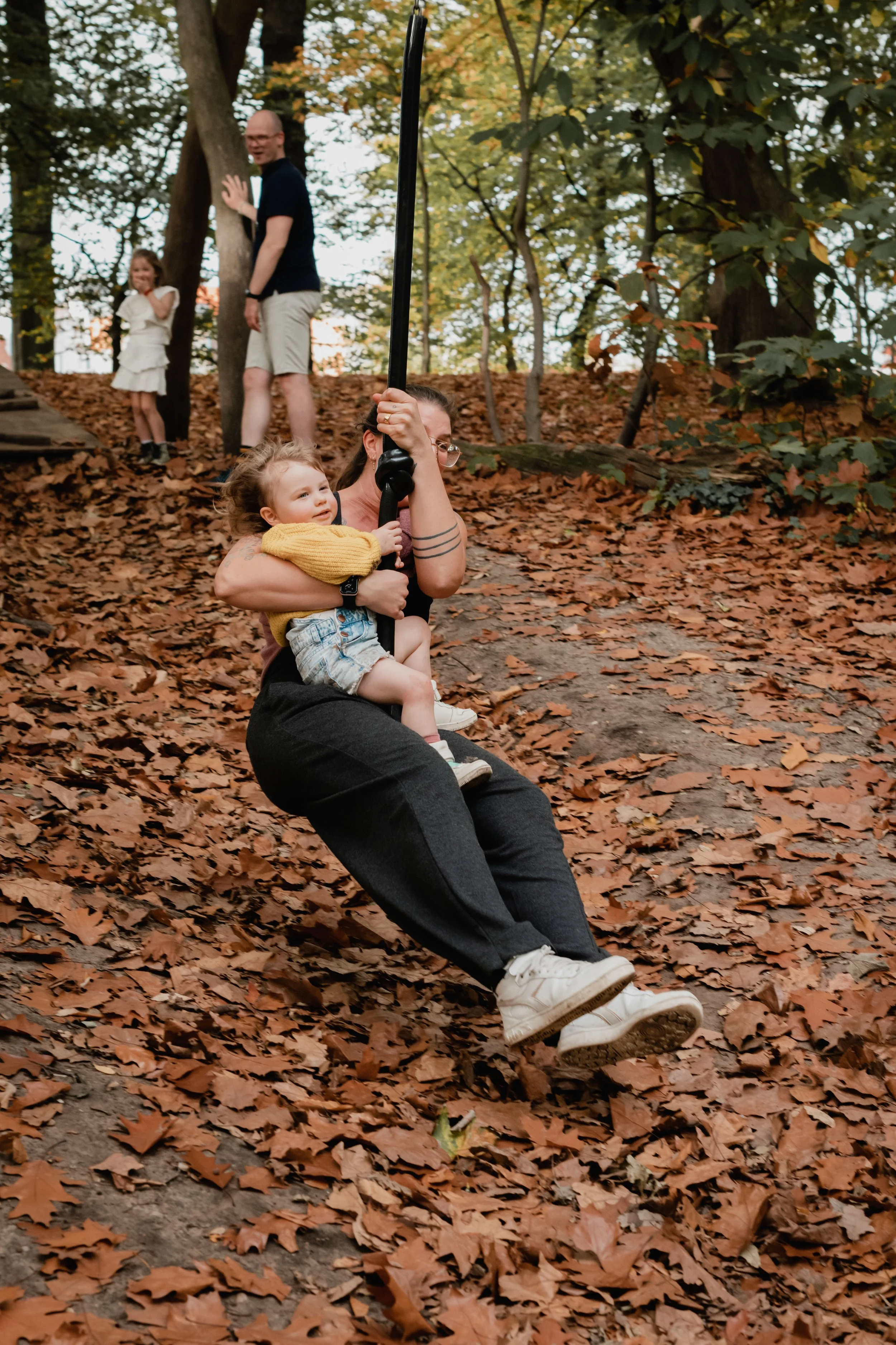 A woman sitting on a hill covered with fallen autumn leaves, holding a child. In the background, a man and a little girl are standing near trees, with the man waving at the camera.