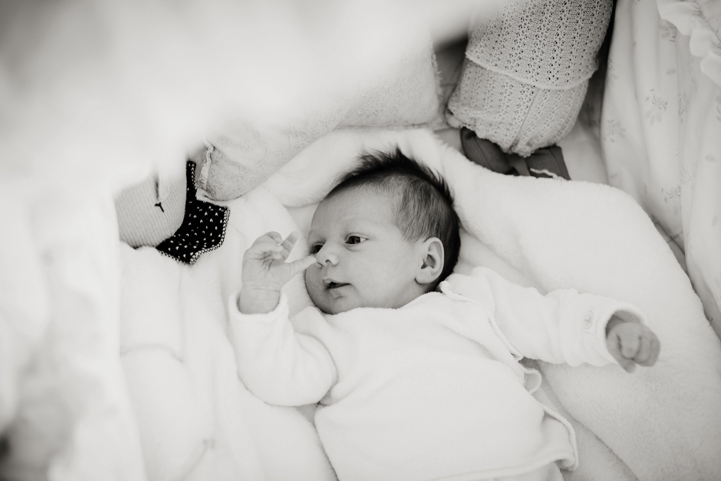 A baby lying on a bed, looking to the side with a hand near its face, surrounded by pillows and blankets.