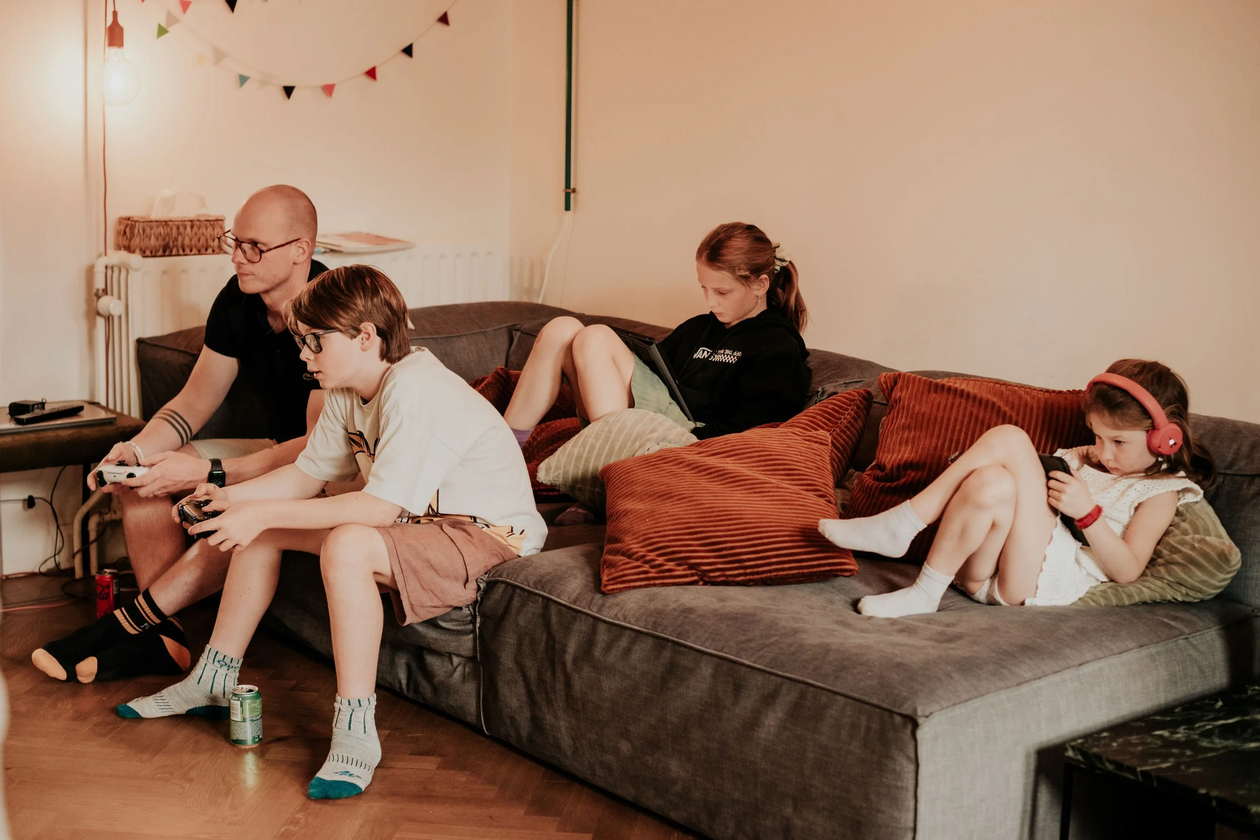 A family of five sitting on a dark gray couch in a living room, playing video games. They are focused on their screens, with two young boys, a girl, and two adult men. The living room has a wooden floor, warm lighting, a small side table with a remot