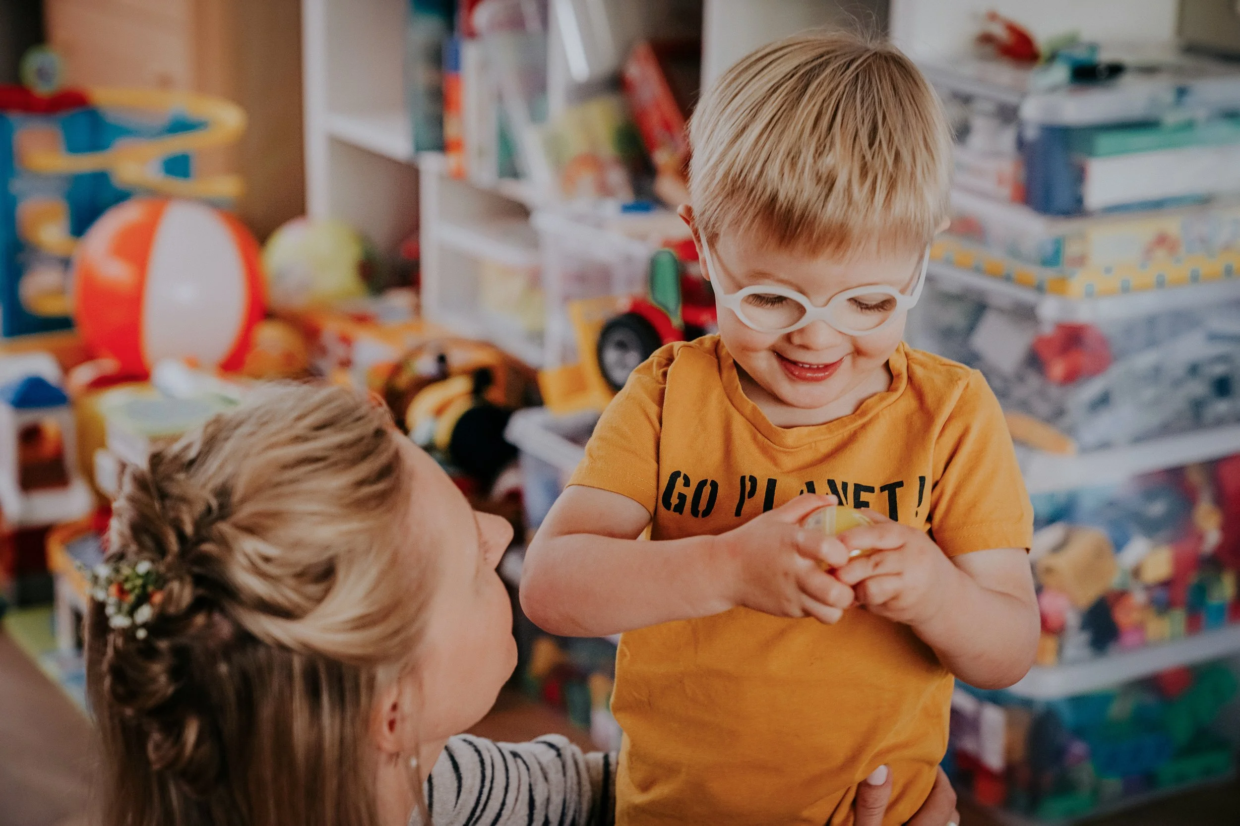 A smiling young boy with blond hair and glasses wearing a yellow shirt with the words 'Go Planet!' is holding a small yellow toy or object. An adult woman with blonde hair styled in a braid is looking at him. The background is filled with shelves of 