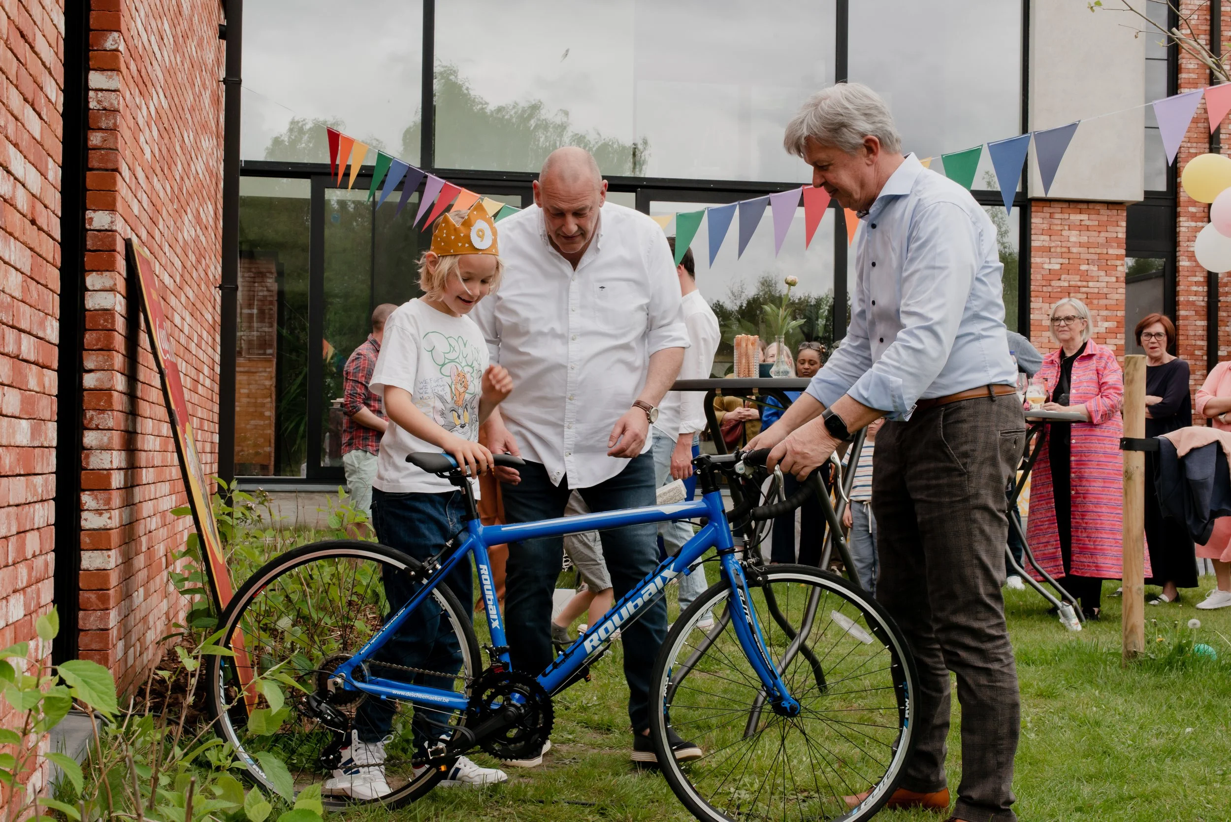 Young girl with a crown learning how to ride a bicycle with two adult men assisting her at a festive outdoor gathering, decorated with colorful bunting and balloons.