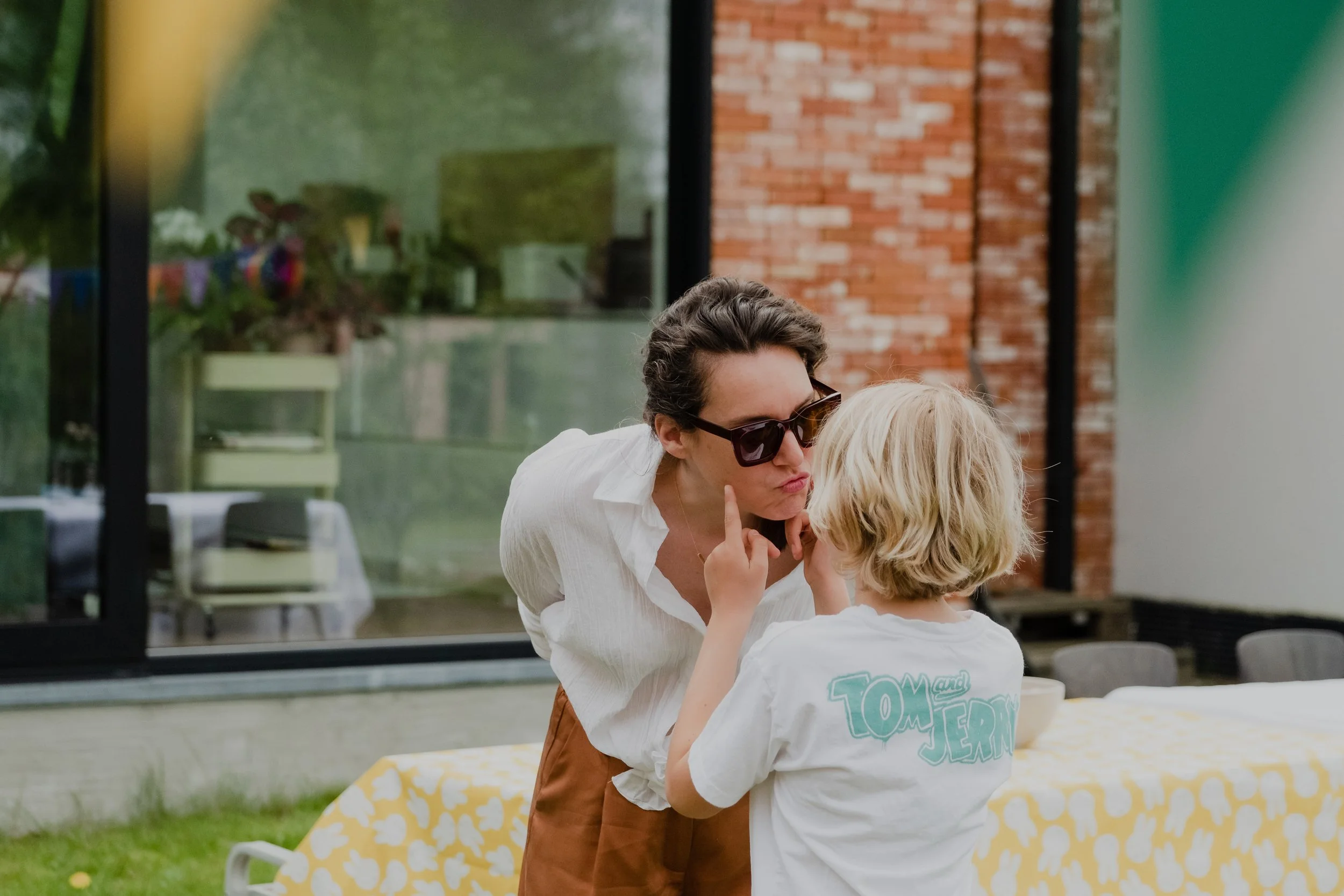 Woman with sunglasses and a child with blonde hair facing each other, outdoors near a table with a yellow patterned tablecloth.