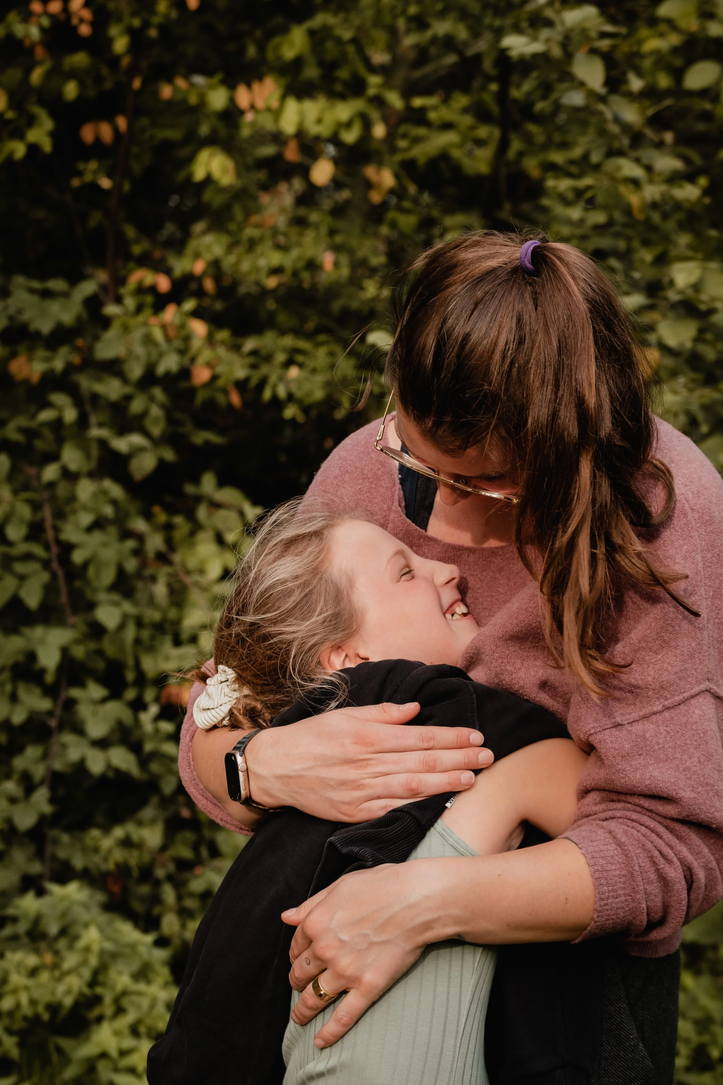 A woman with brown hair, glasses, and a purple hair tie, hugging a young girl with blonde hair tied back with a white scrunchie, smiling happily, outside at night with greenery in the background.