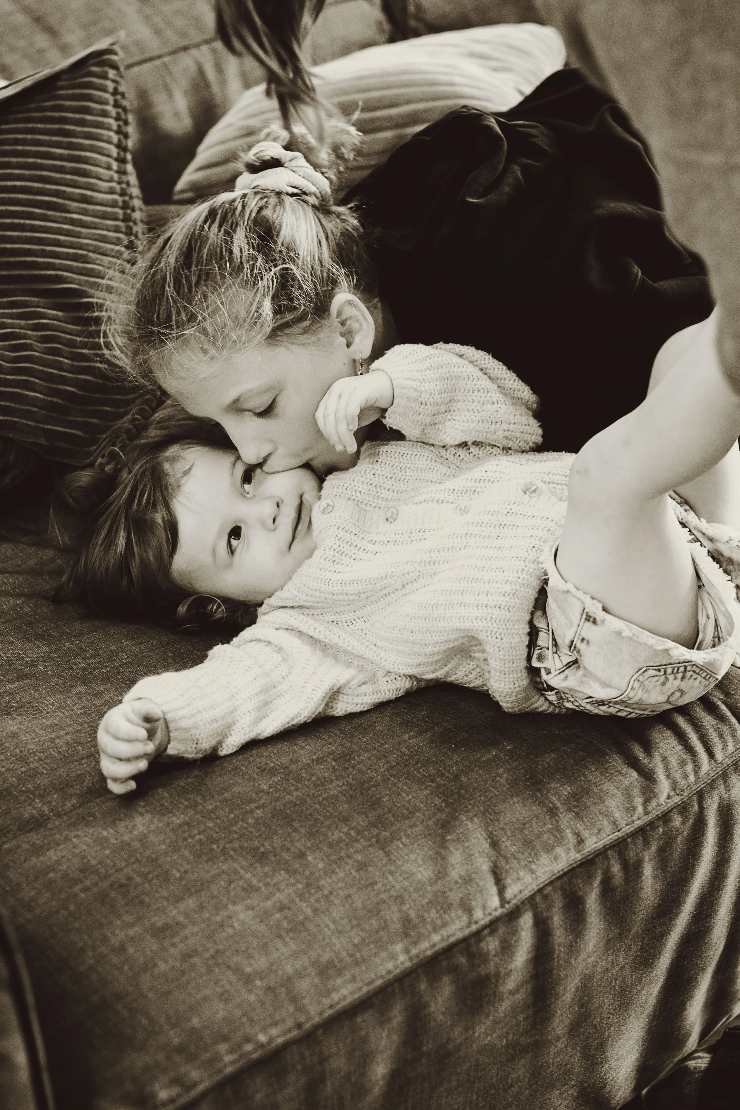 A woman and a young girl are lying on a couch, sharing an affectionate moment. The woman kisses the girl on the forehead while the girl looks up at her with a smile. The girl is wearing a knitted sweater and shorts, and the woman has her hair tied ba