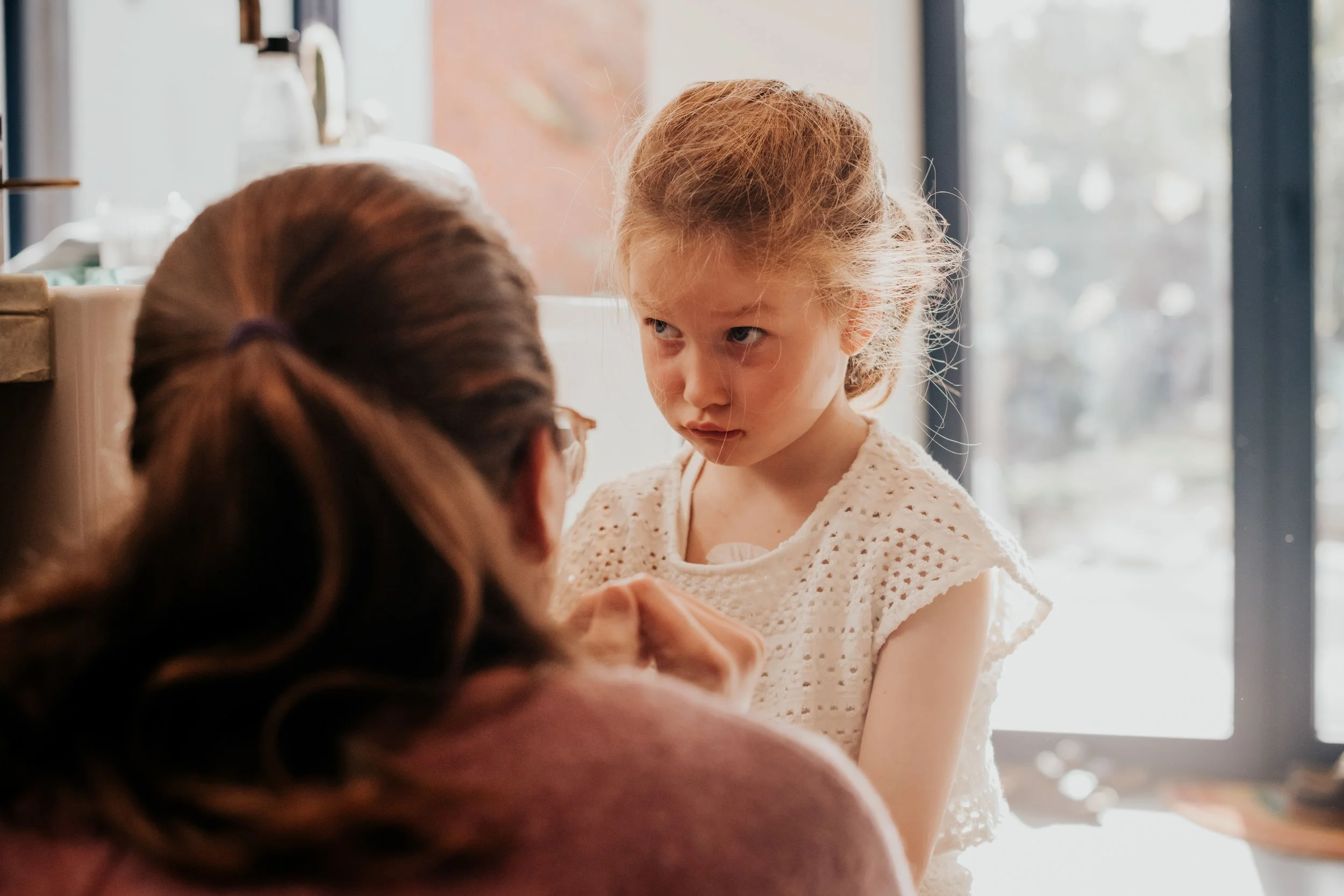 A young girl with curly red hair and blue eyes looking intently at a woman with brown hair tied in a ponytail, both inside a room with large windows and natural light.