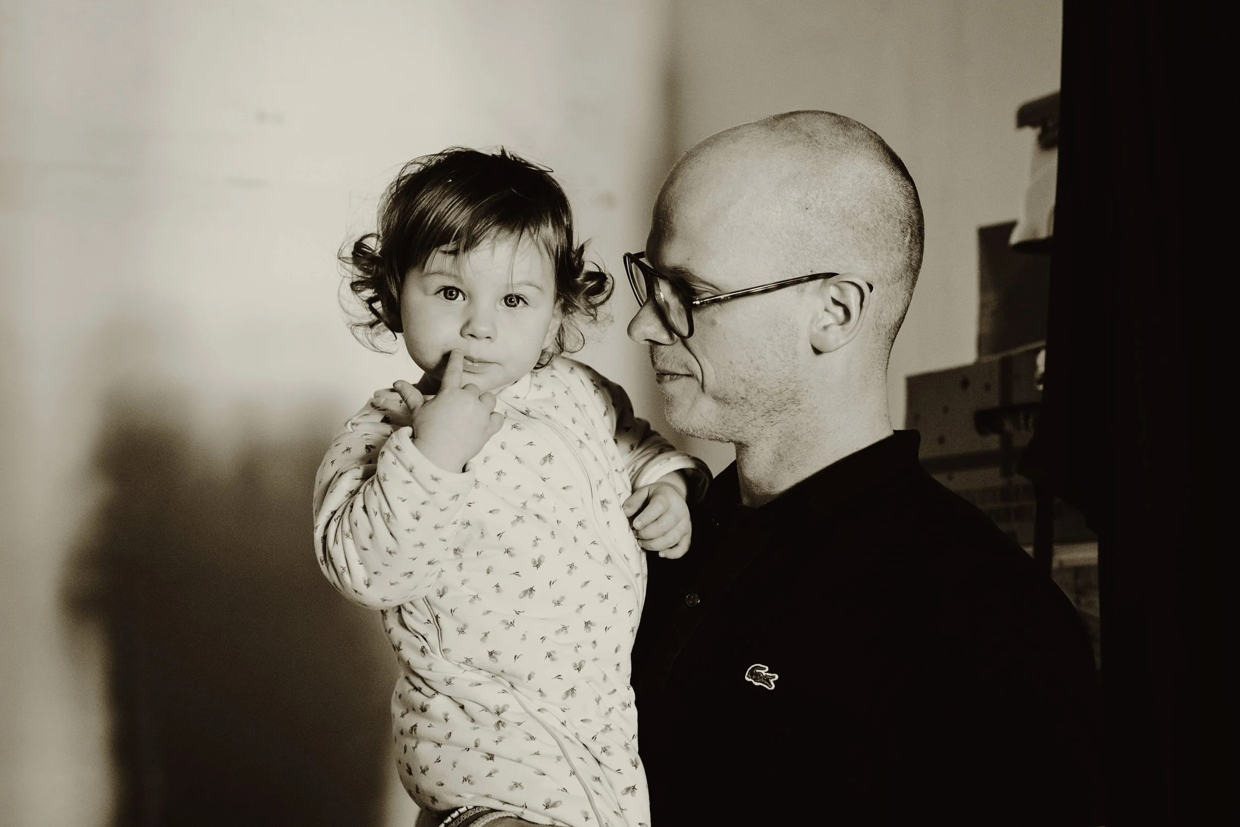 A man with glasses holding a young girl, who has curly hair and is touching her finger to her lips, in a room with a plain background and industrial shelves.