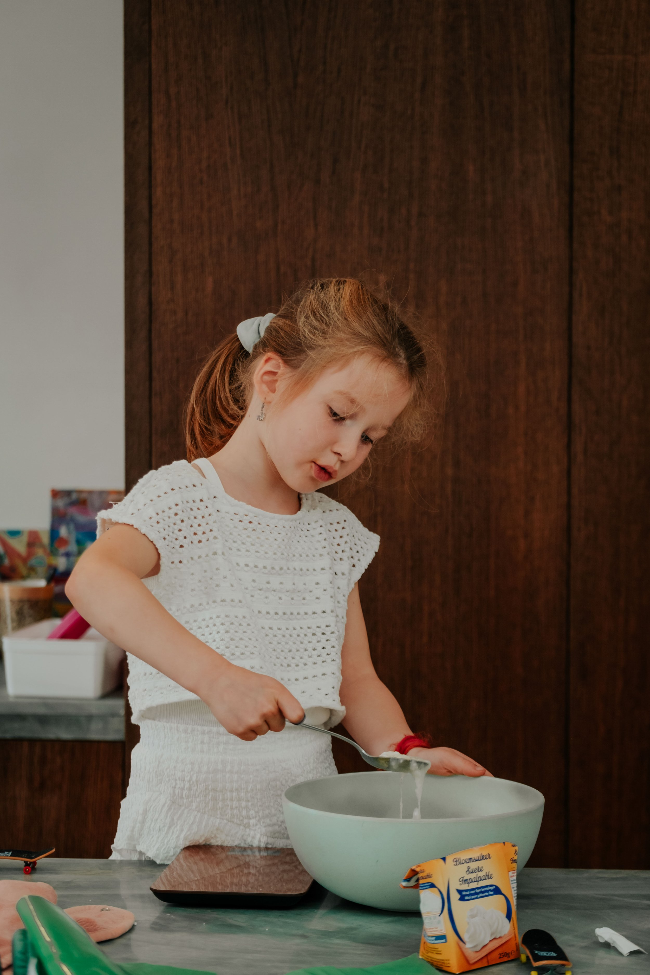 A young girl with red hair wearing a white crochet top, standing at a kitchen counter, scooping whipped cream from a container into a large bowl.
