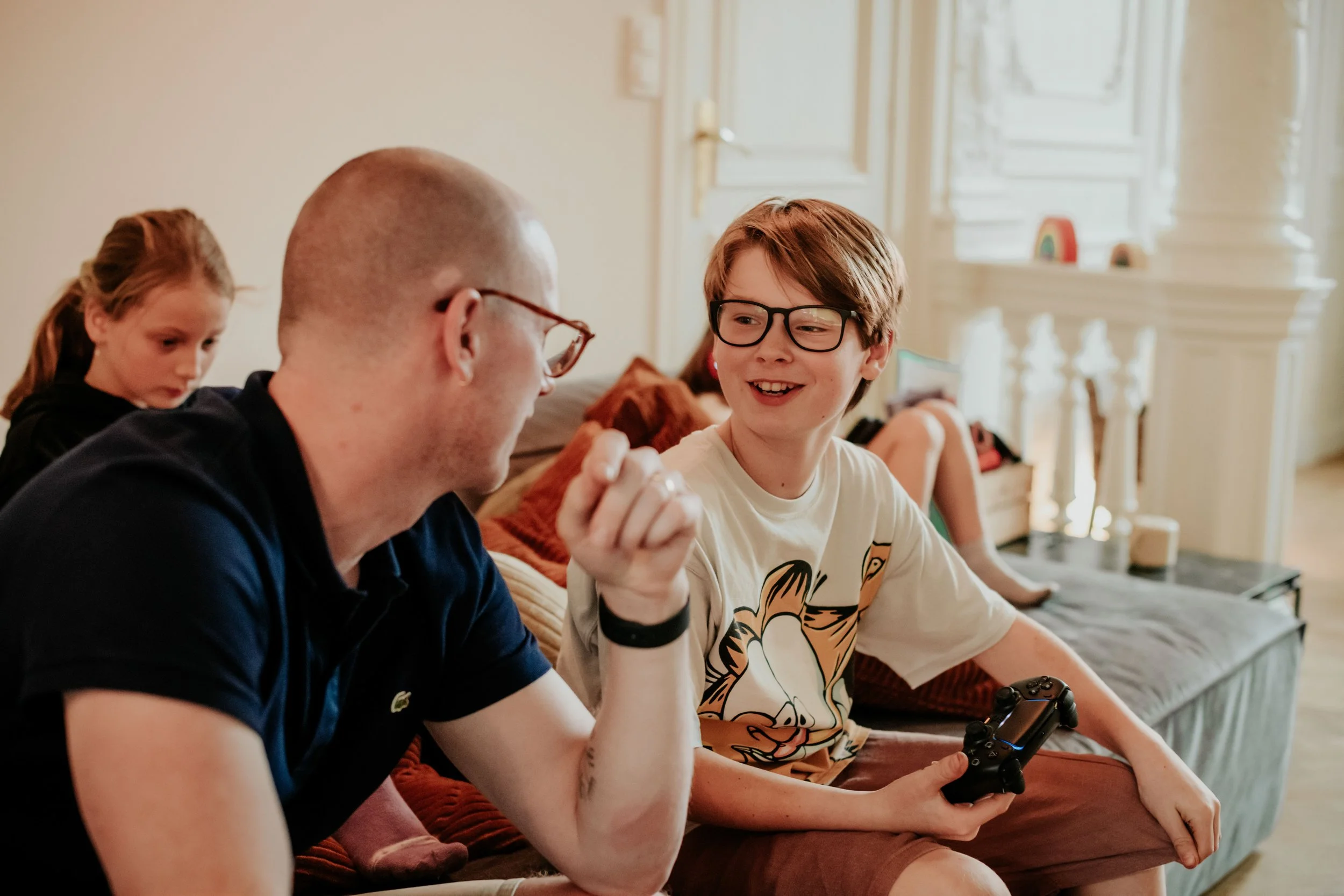 A group of people sitting on a couch indoors playing video games and talking, with a young boy in glasses holding a game controller smiling.