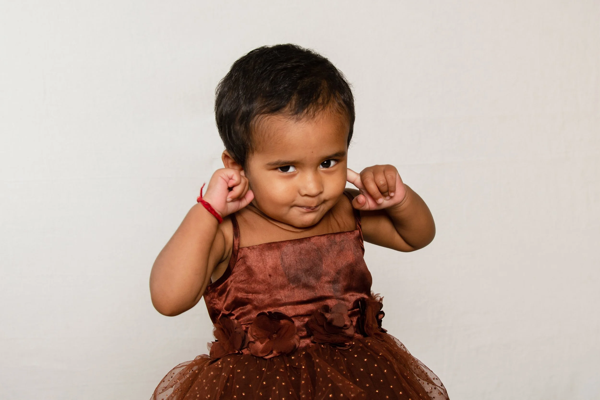 A young girl with dark hair and brown skin wearing a brown dress with floral embellishments and a red bracelet, standing against a plain white background, covering her ears with her fingers.