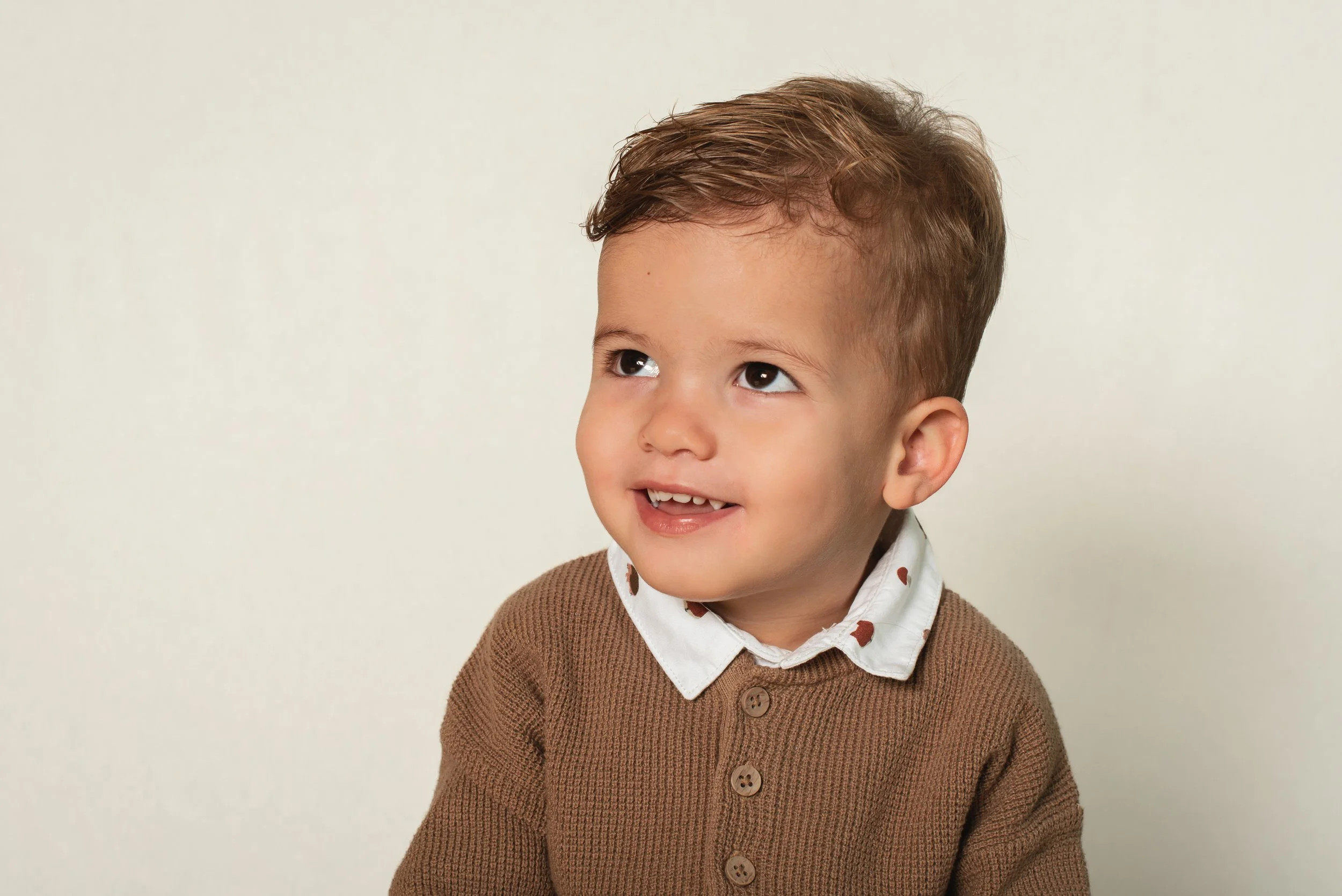 A young boy with brown hair wearing a white shirt with a brown heart pattern and a brown sweater, smiling and looking up, against a plain light background.