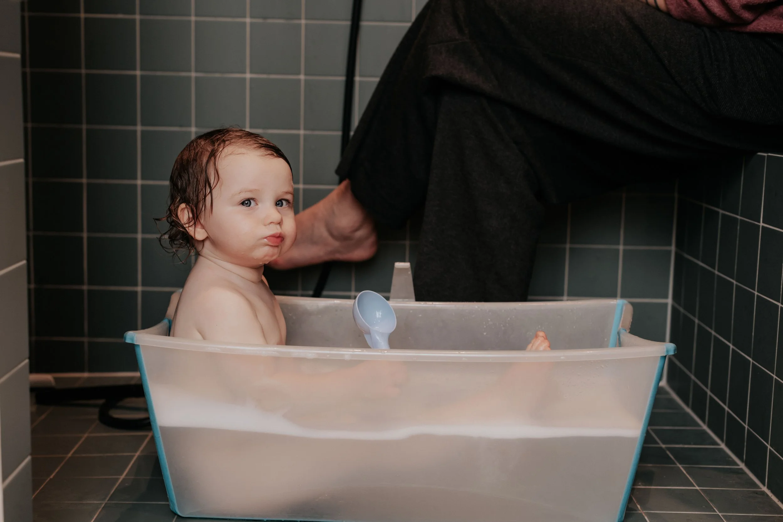 A young child with wet hair sitting in a bathtub with a surprised or curious expression while a person outside the tub bathes them. The background features dark tiled bathroom walls.