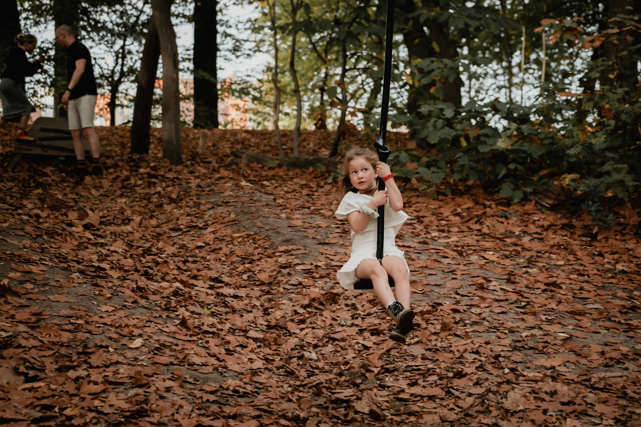 Young girl in a white dress on a swing in a wooded park with fallen leaves.