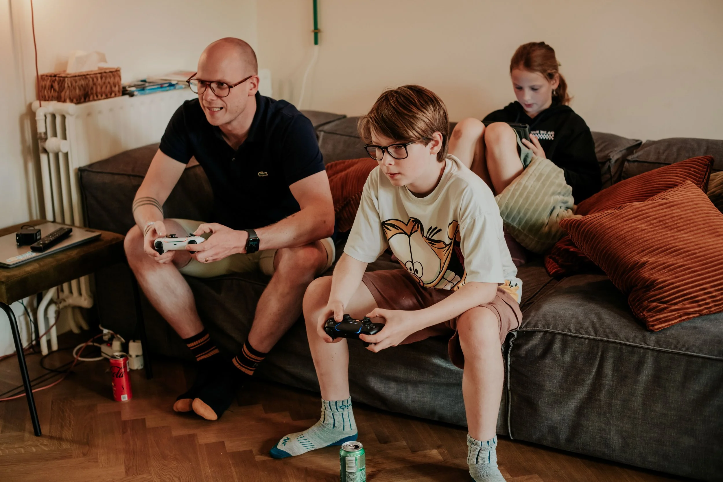 A man and two children sitting on a brown couch playing video games with controllers. The man is wearing glasses and a black shirt. The boy in front is wearing glasses, a beige shirt with a character design, and brown shorts. The girl in back is usin