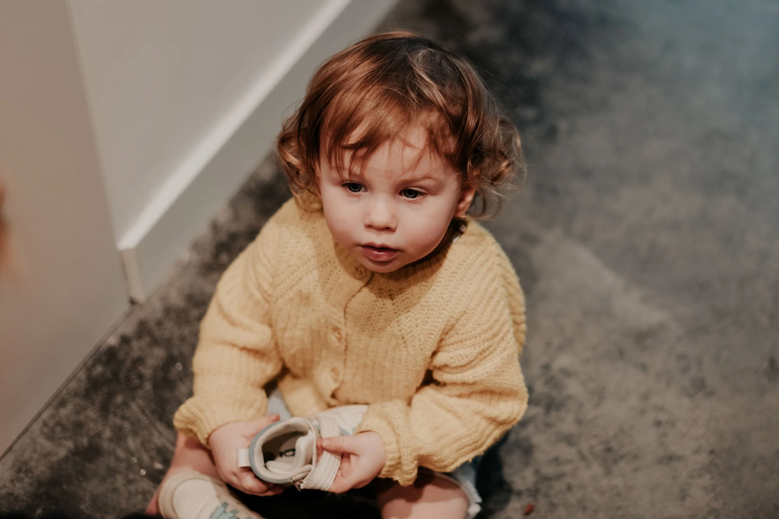 A young boy with curly brown hair and blue eyes, wearing a beige knit sweater, sitting on a dark, textured carpeted floor, holding a pair of beige sneakers.