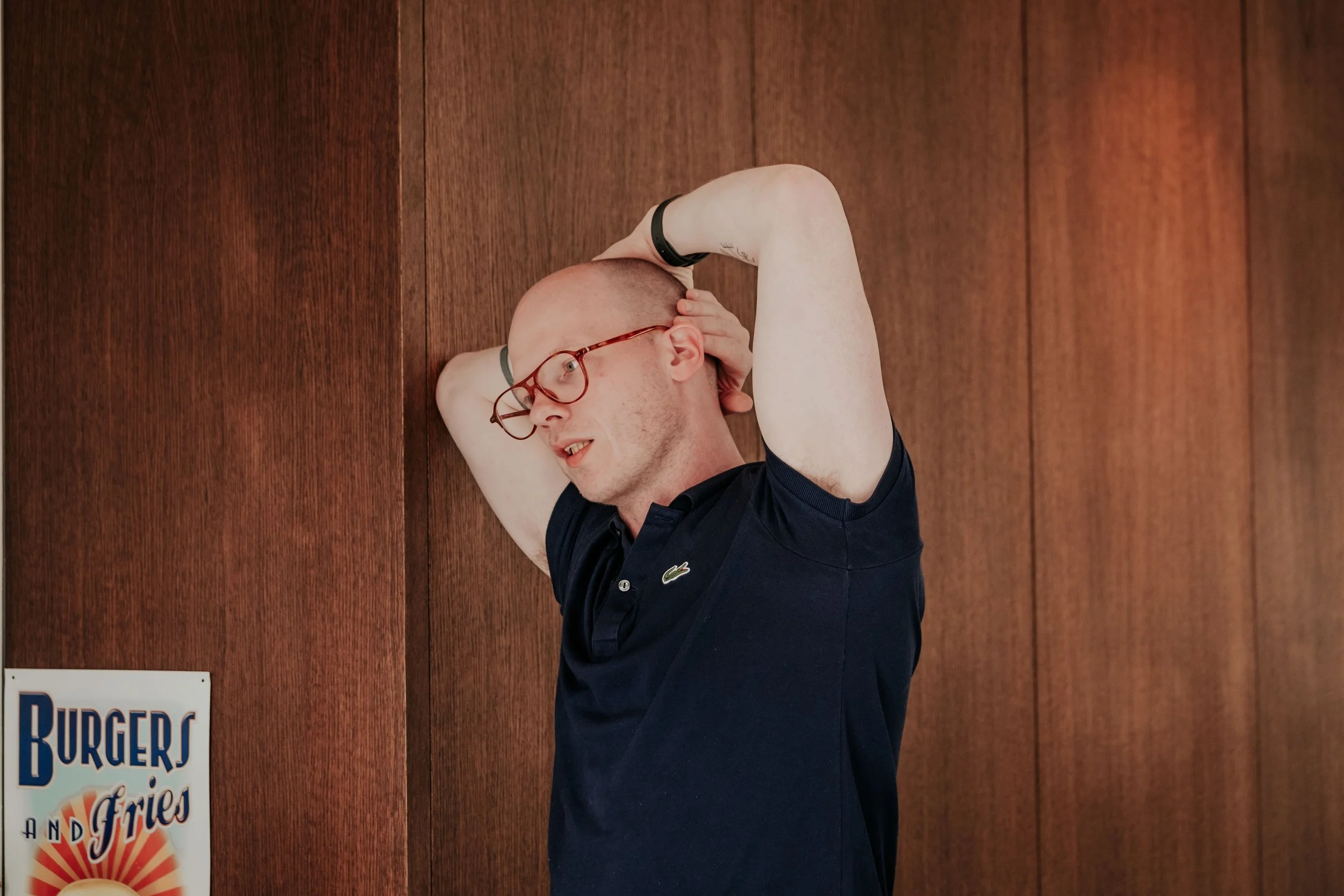 A bald man wearing red glasses and a black wristband, dressed in a navy blue polo shirt, stands with his eyes closed and one hand on his head against a wood-paneled wall.