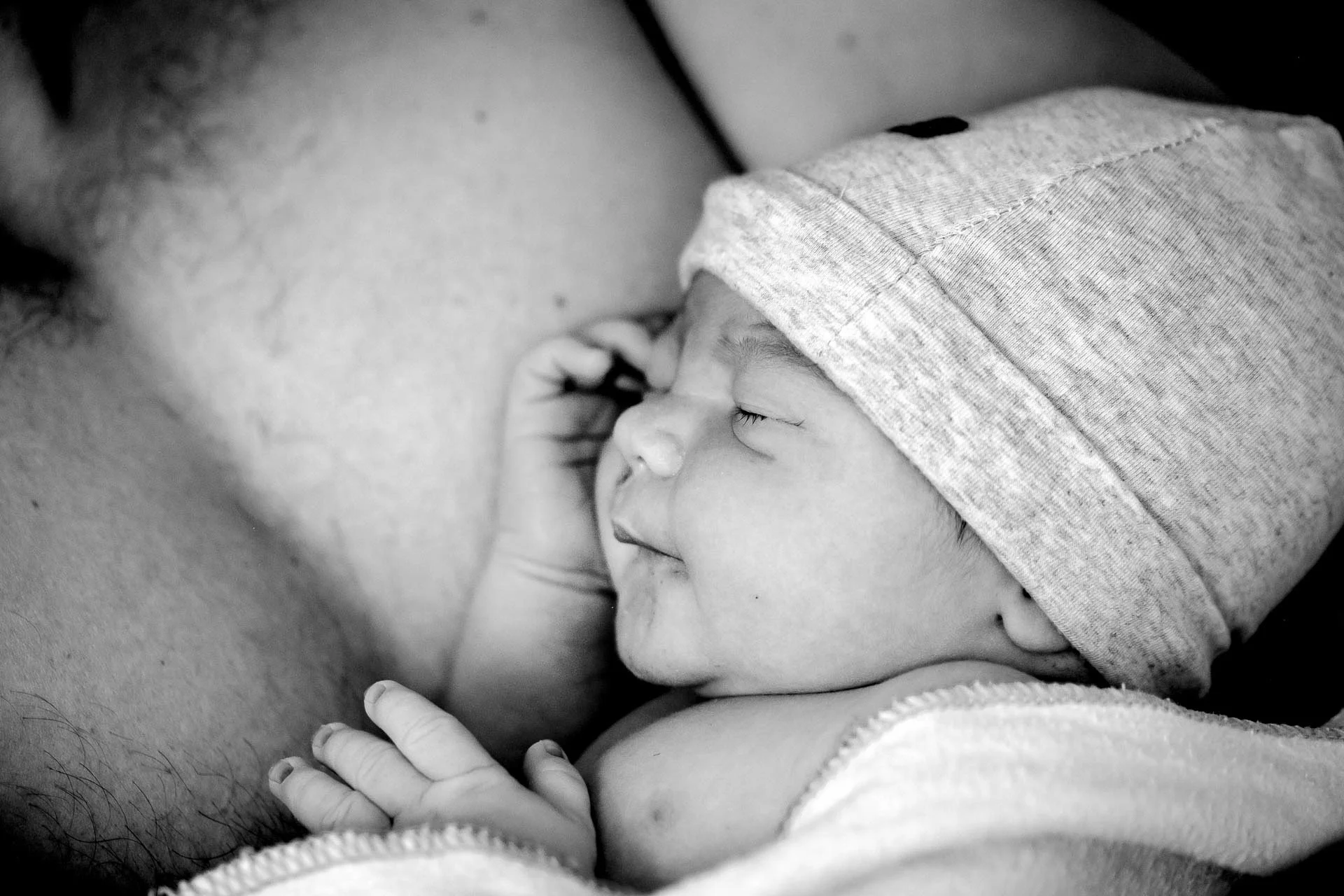 Black and white photo of a father holding his newborn baby close to his face, with the baby wearing a hoodie and sleeping peacefully.