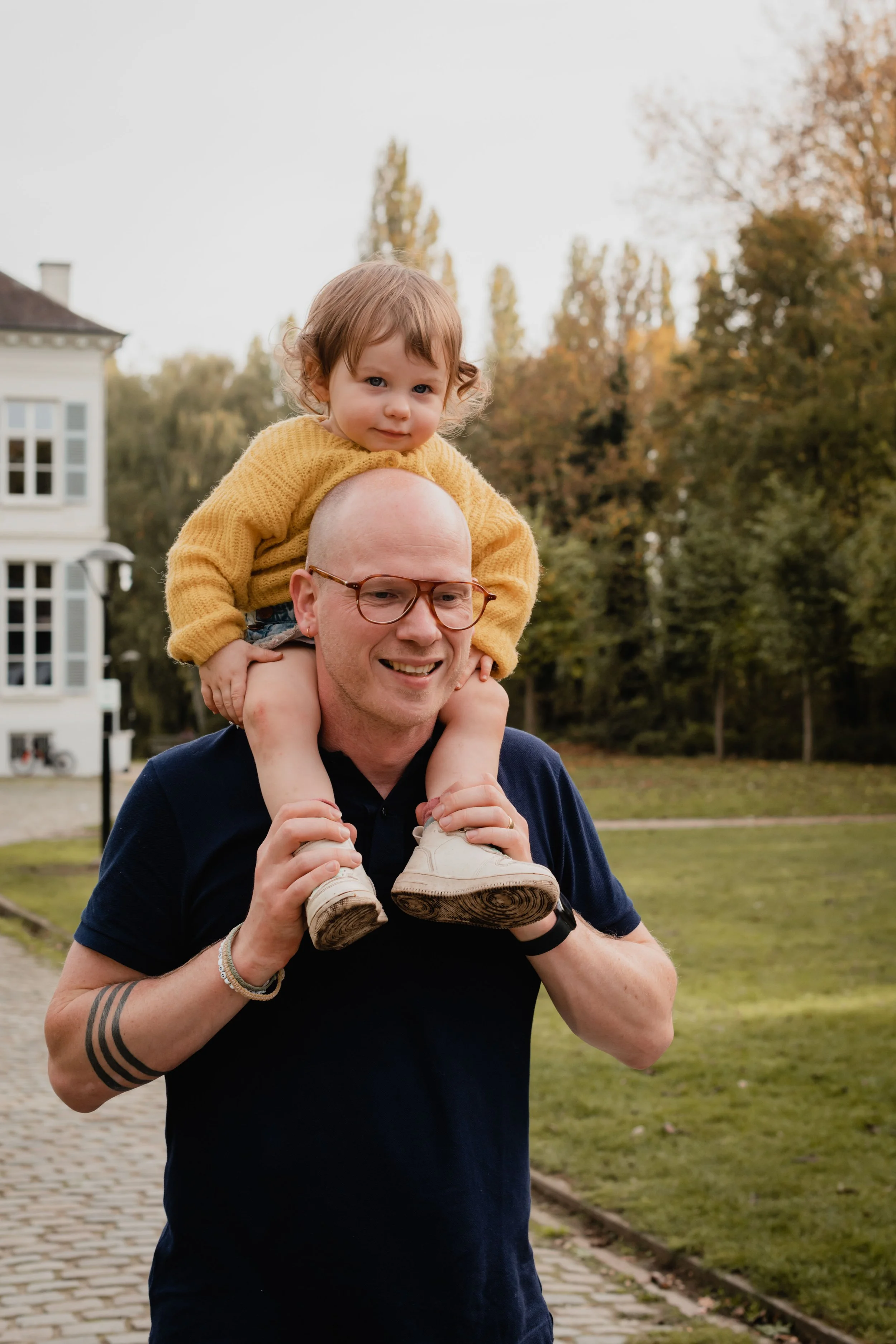 A man wearing glasses and a black shirt is smiling while carrying a young child on his shoulders outdoors with trees and a building in the background.