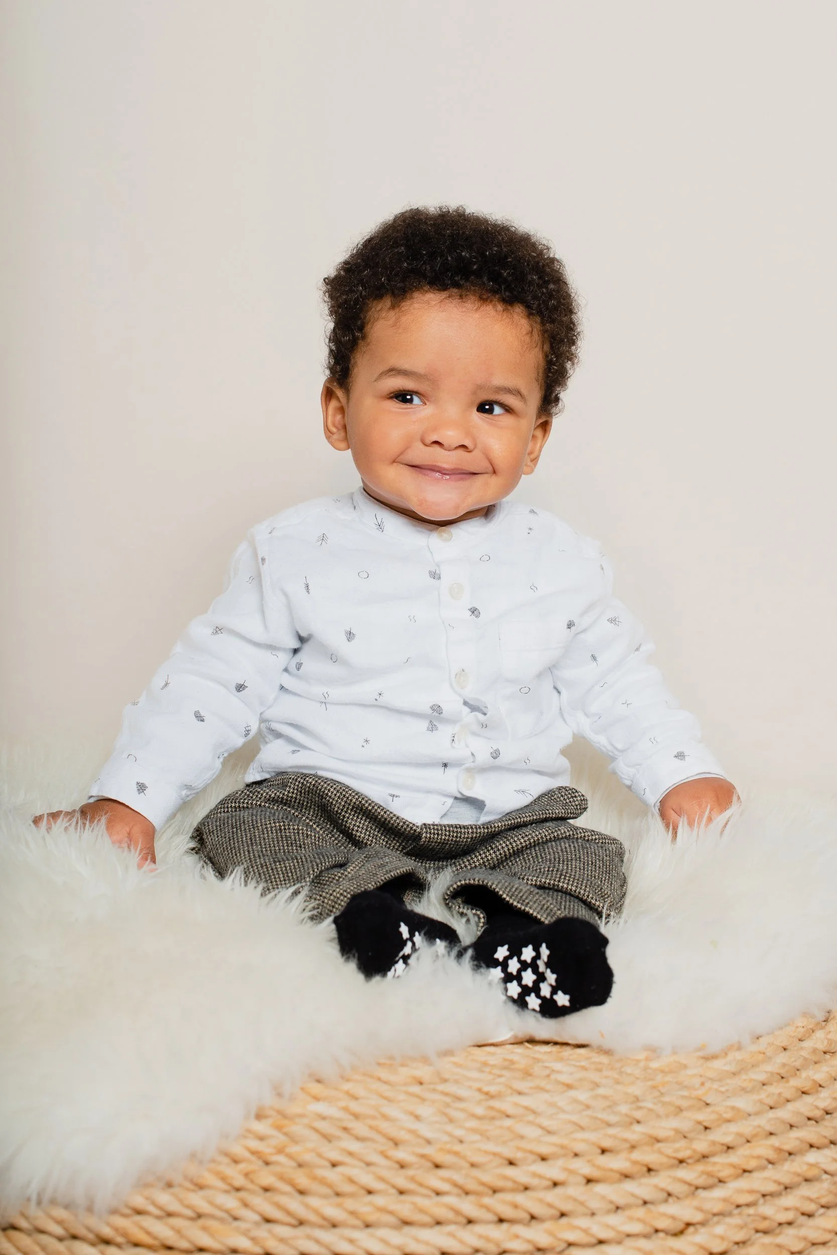 A young child with curly hair smiling while sitting on a fluffy white rug in front of a plain white wall, wearing a white button-up shirt with small patterns, gray plaid pants, and black socks with white stars.