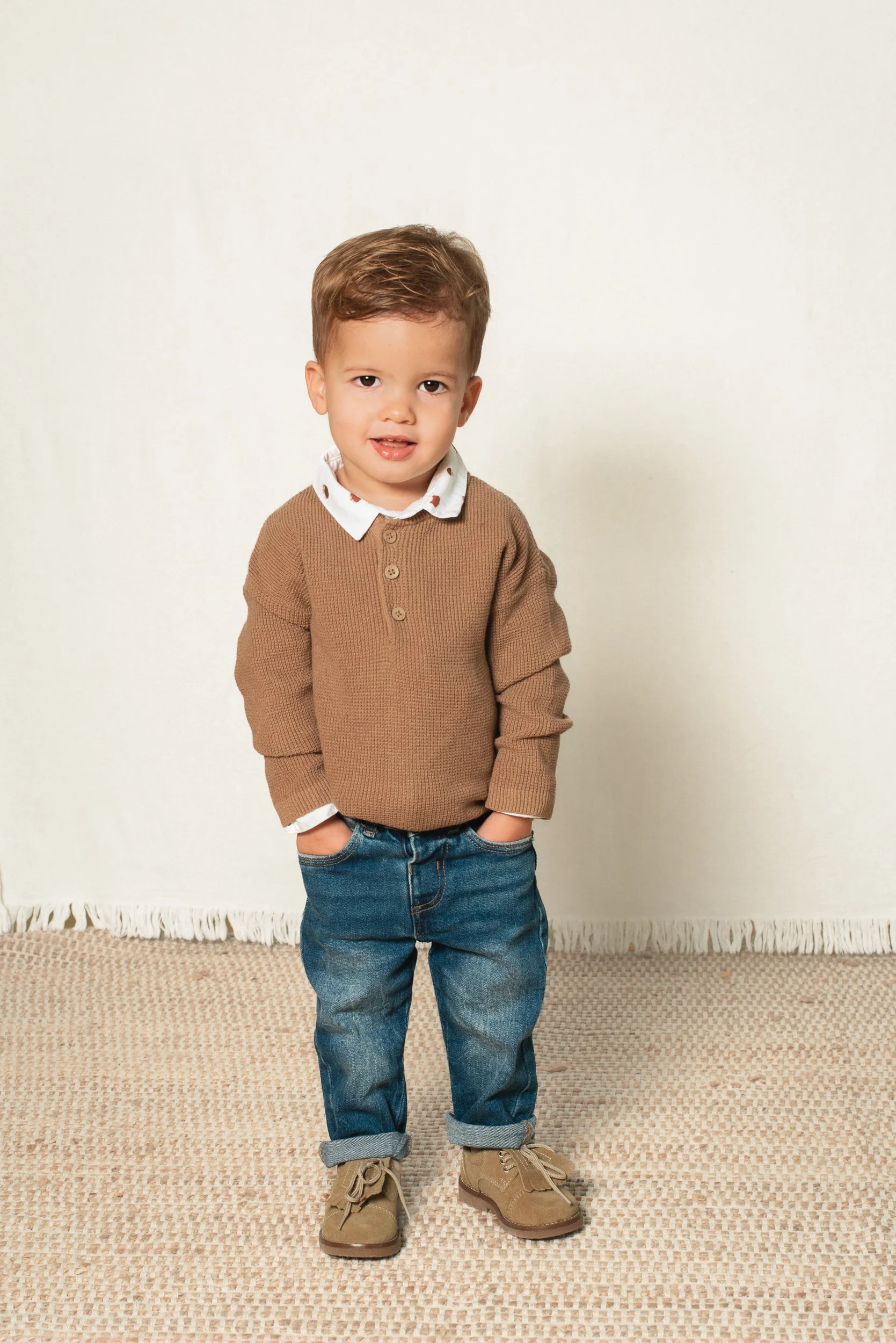A young boy with light skin and short brown hair stands indoors against a plain white wall, facing the camera with a slight smile, dressed in a brown sweater, jeans, and tan boots.