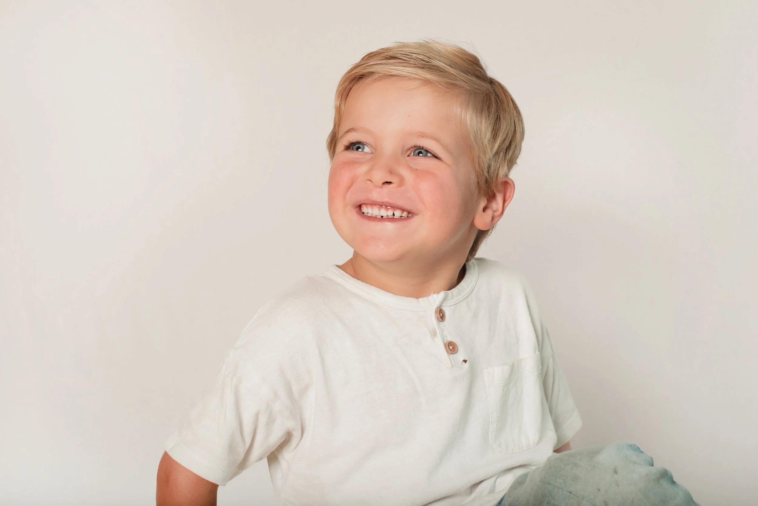 A young boy with blonde hair and blue eyes smiling, wearing a cream-colored shirt with buttons, against a plain light background.