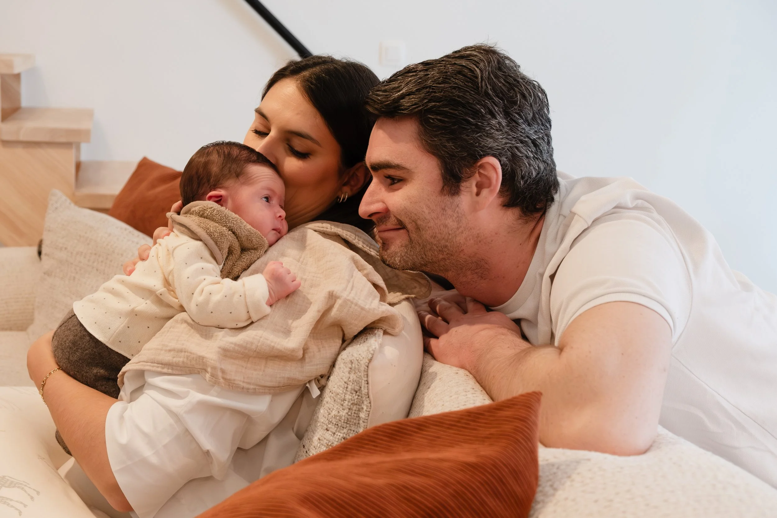 Family of three—mother, father, and newborn—lying on a bed, sharing a tender moment and gazing at the baby.