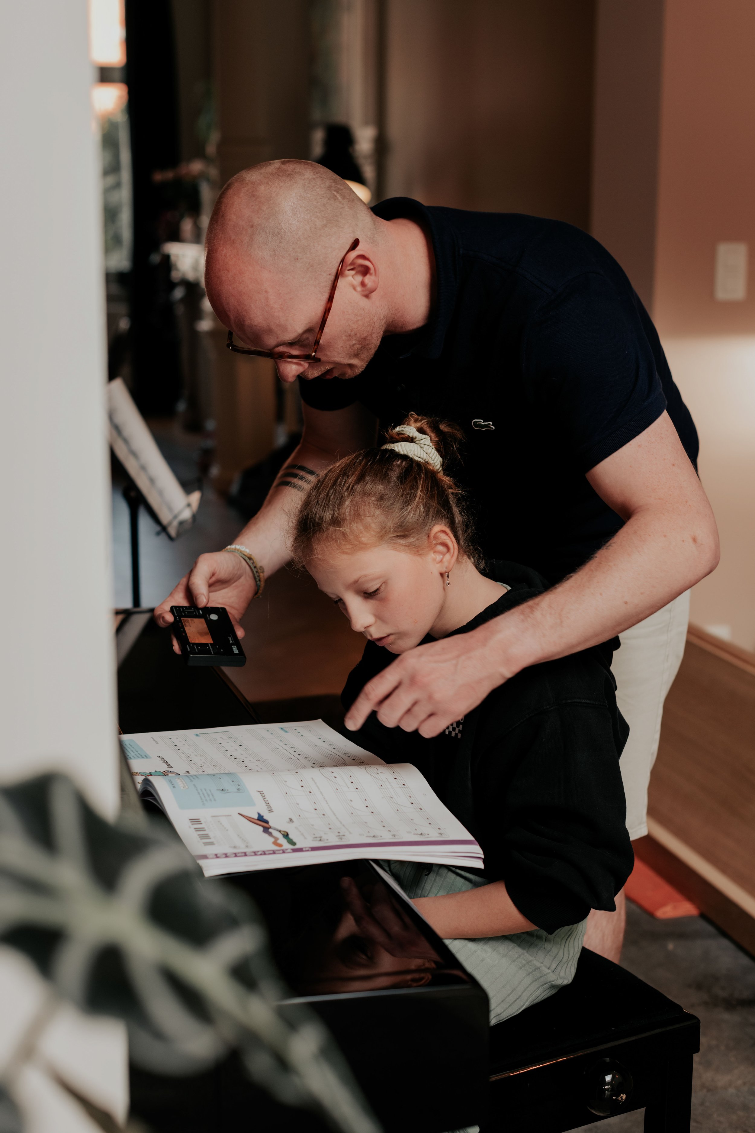 A man and a young girl are looking at a book on a piano, with the man leaning over and helping the girl read.