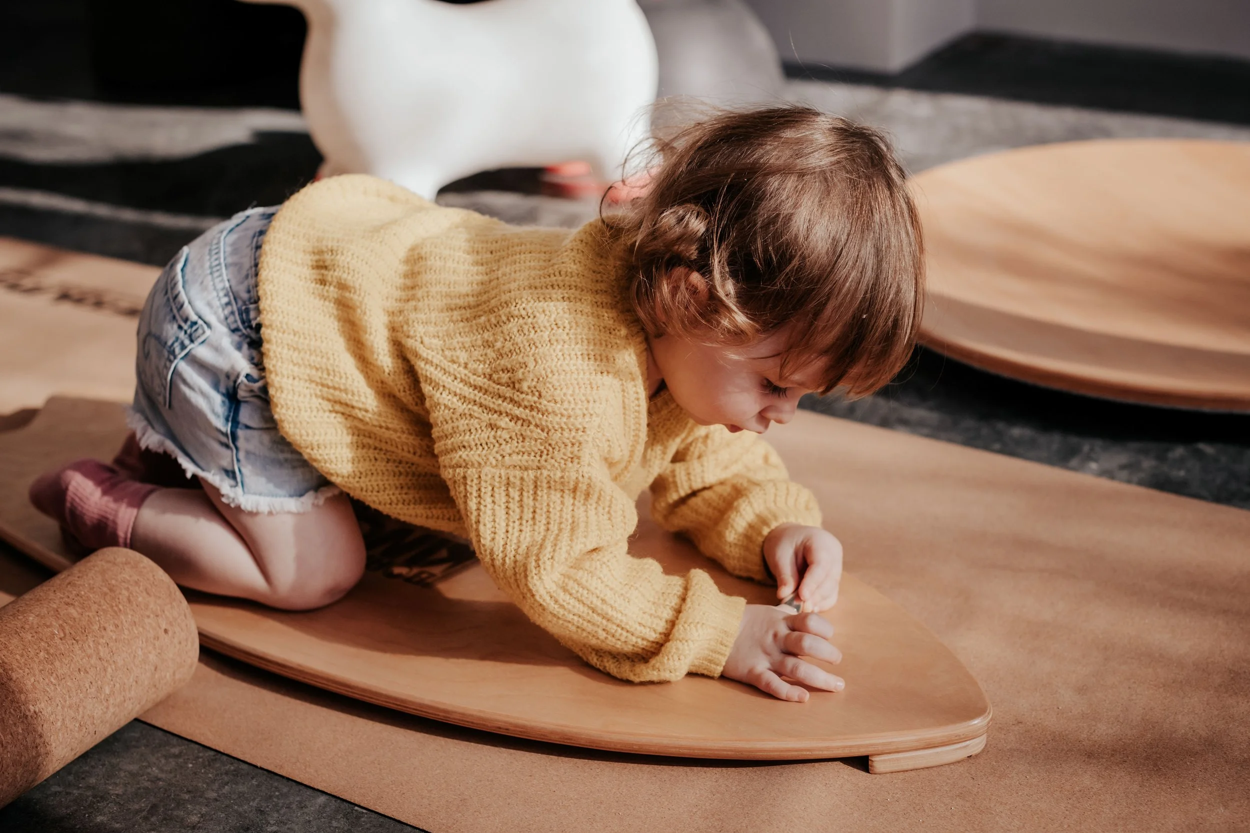 A young child with brown hair, wearing a yellow sweater and shorts, is crawling on a wooden surface. There are large round objects, resembling handles or decorative items, and a white sculpture of a duck in the background.