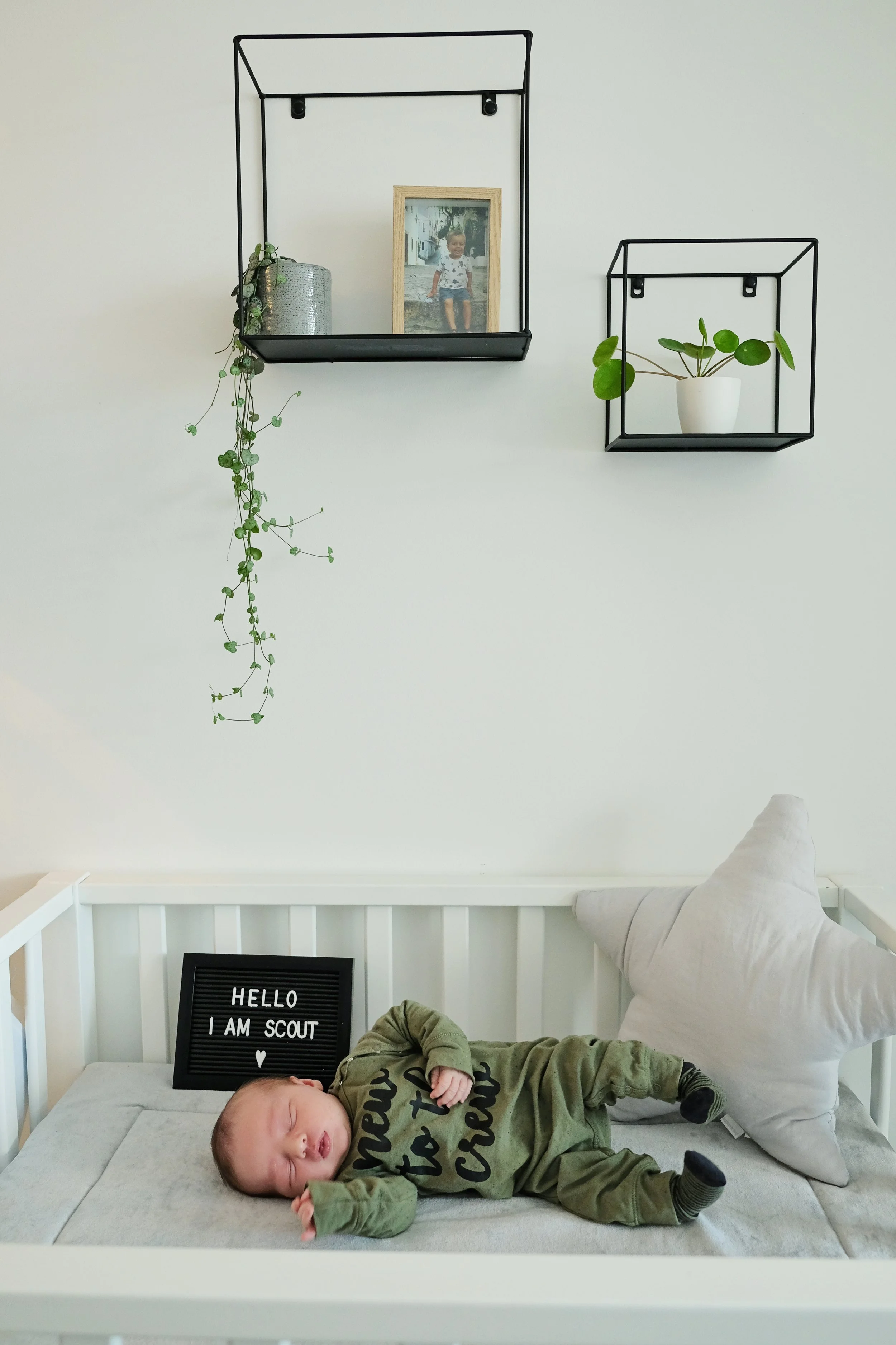 A baby sleeping on a bed with a white frame, next to a large gray pillow shaped like a star. The baby is dressed in green pajamas. Behind the baby, there's a black letter board with white text that reads, "HELLO I AM SCOUT". On the wall above the bed