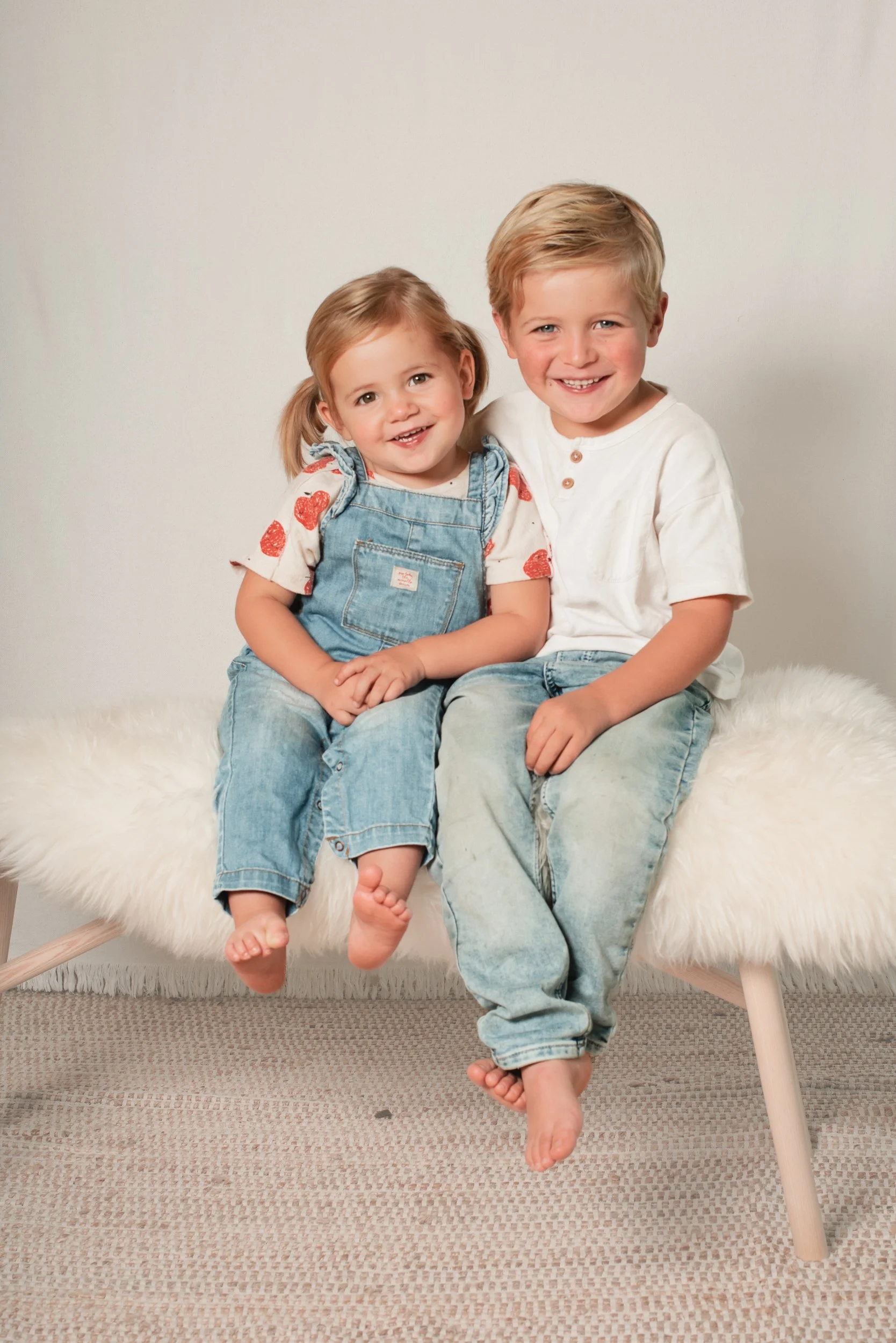 Two young children, a girl and a boy, sitting on a white furry bench against a plain background, smiling at the camera.