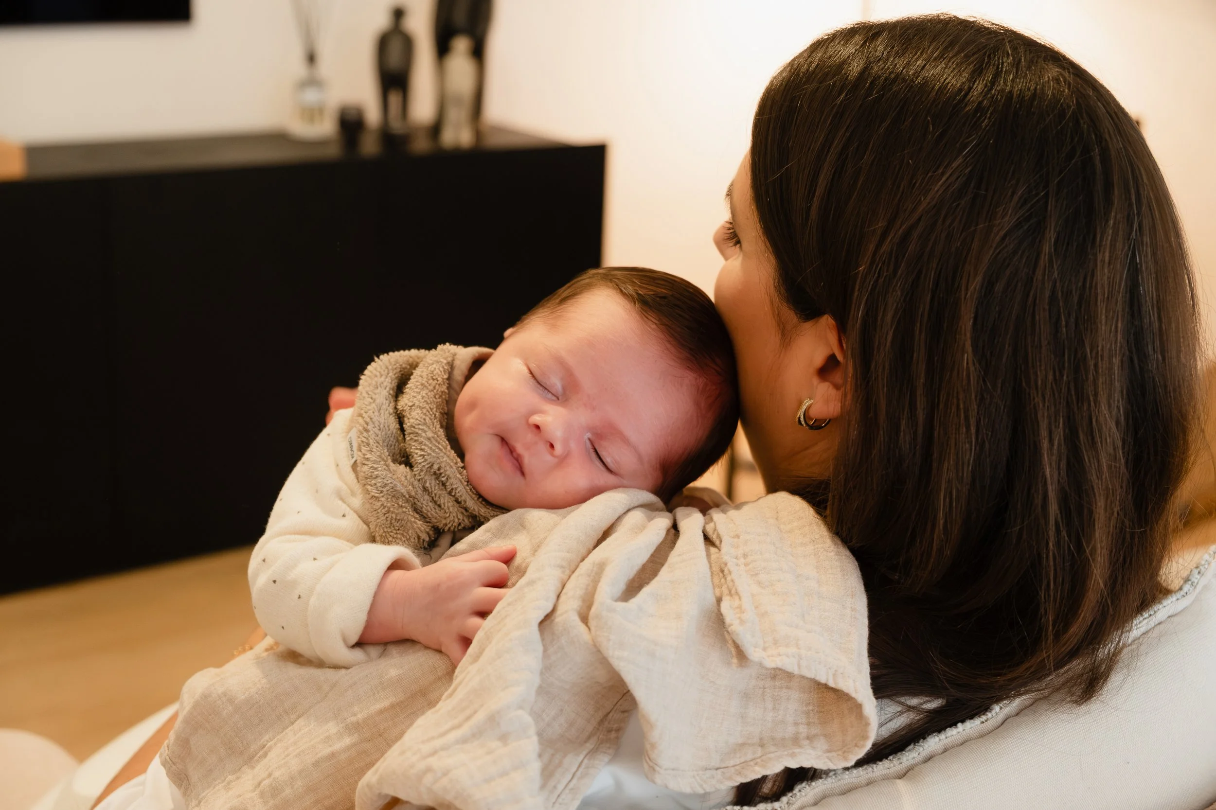A woman with dark brown hair holding and kissing a sleeping baby with light skin and brown hair in an indoor setting.