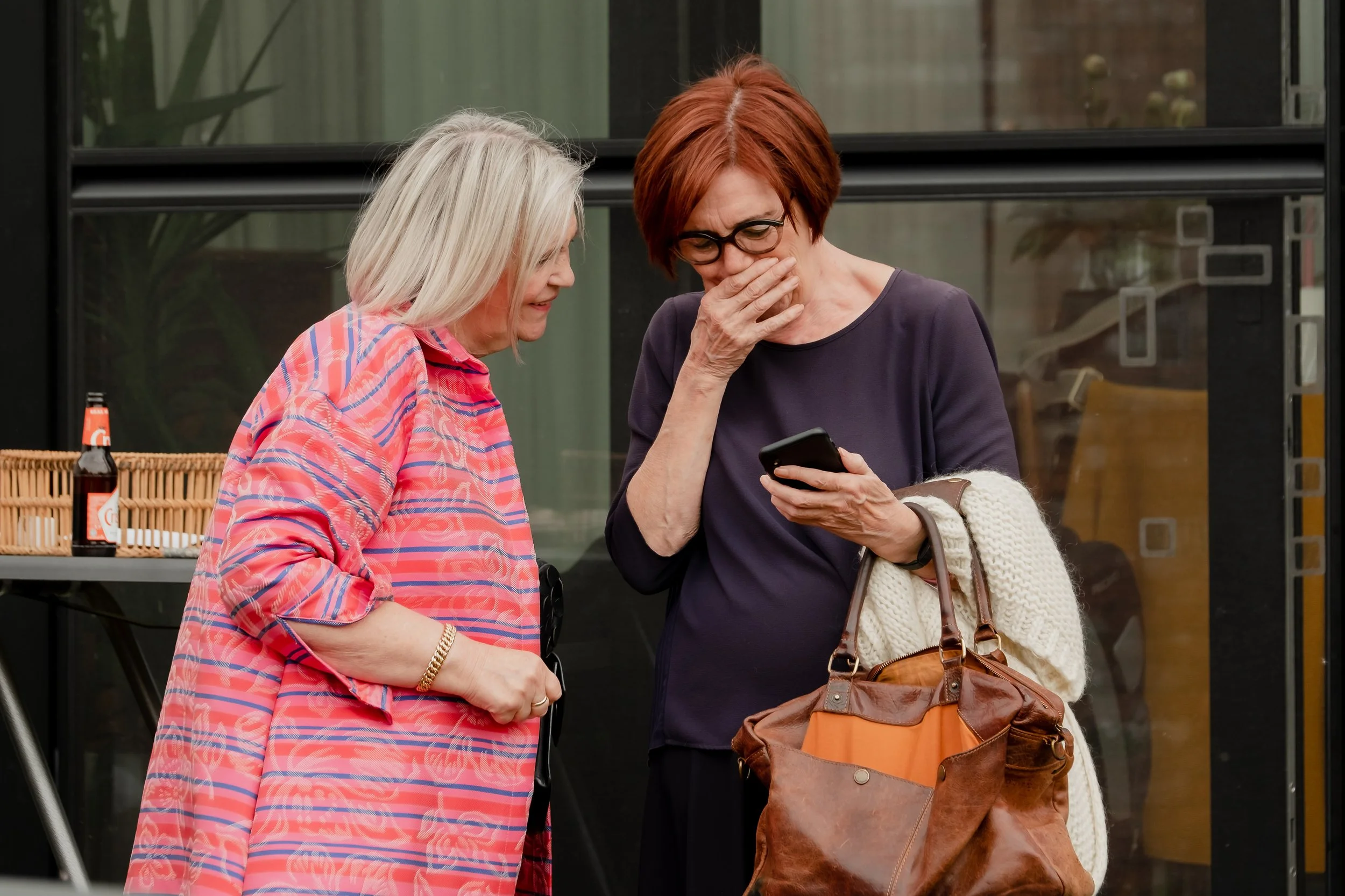 Two women are looking at a smartphone, one with blonde hair wearing a pink patterned shirt and the other with red hair wearing glasses and a dark shirt, in an outdoor setting.