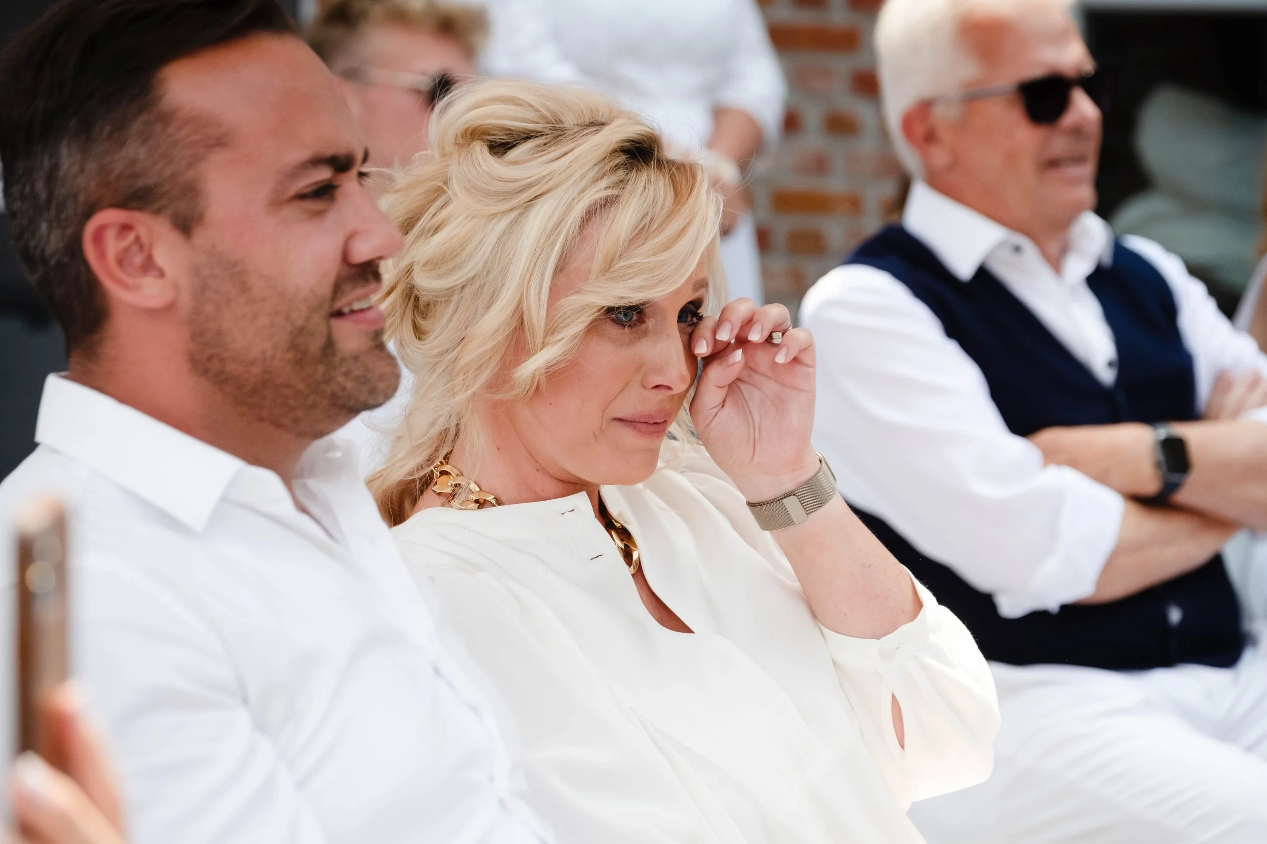 People sitting at a table, including a woman adjusting her glasses and men wearing sunglasses, in an indoor setting with brick walls.