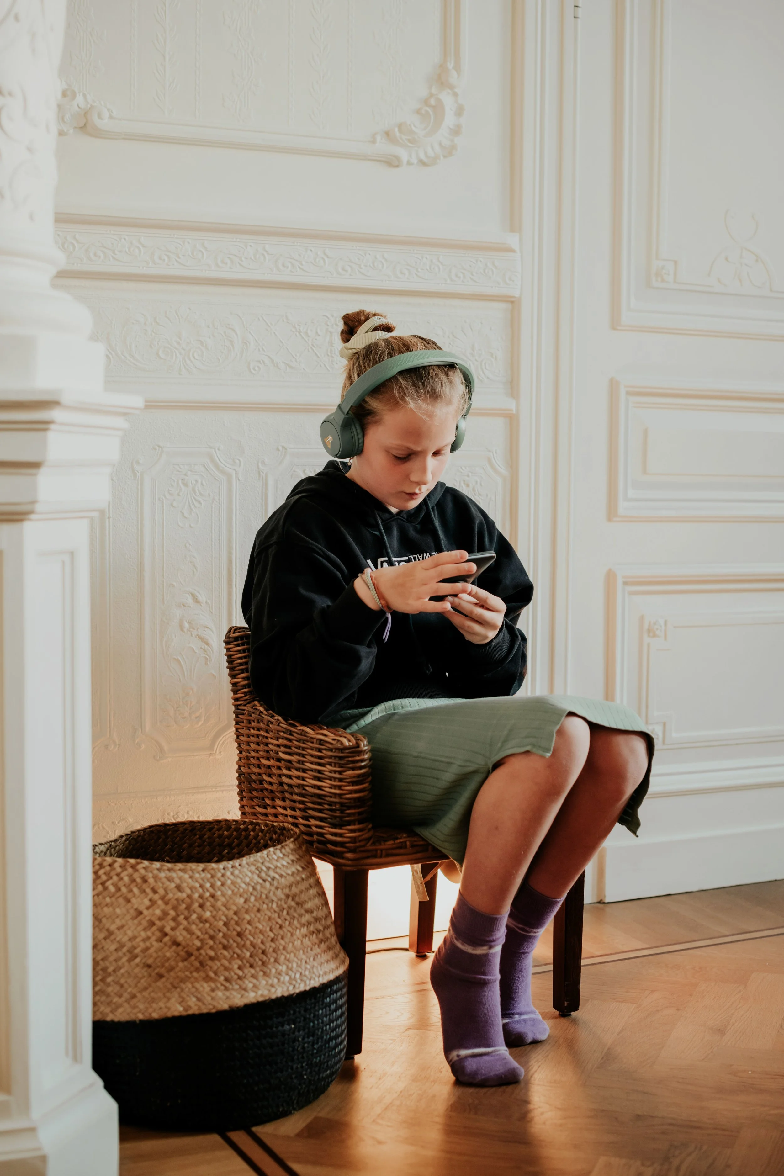 A young girl with a bun hairstyle, wearing a black hoodie, green skirt, and purple socks, sitting on a woven chair and looking at her phone in a room with ornate white wall paneling, hardwood flooring, and a large woven basket nearby.