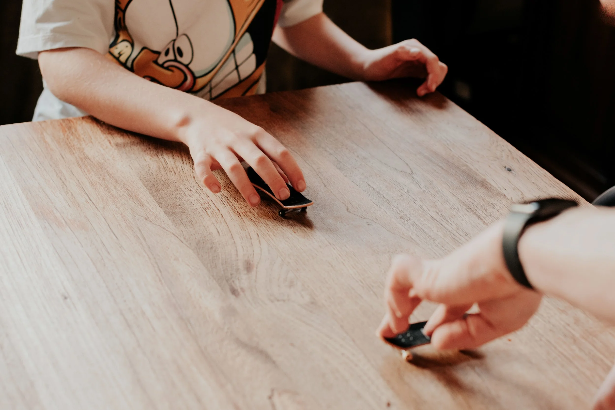 A child in a cartoon character T-shirt is playing with a small skateboard on a wooden table.
