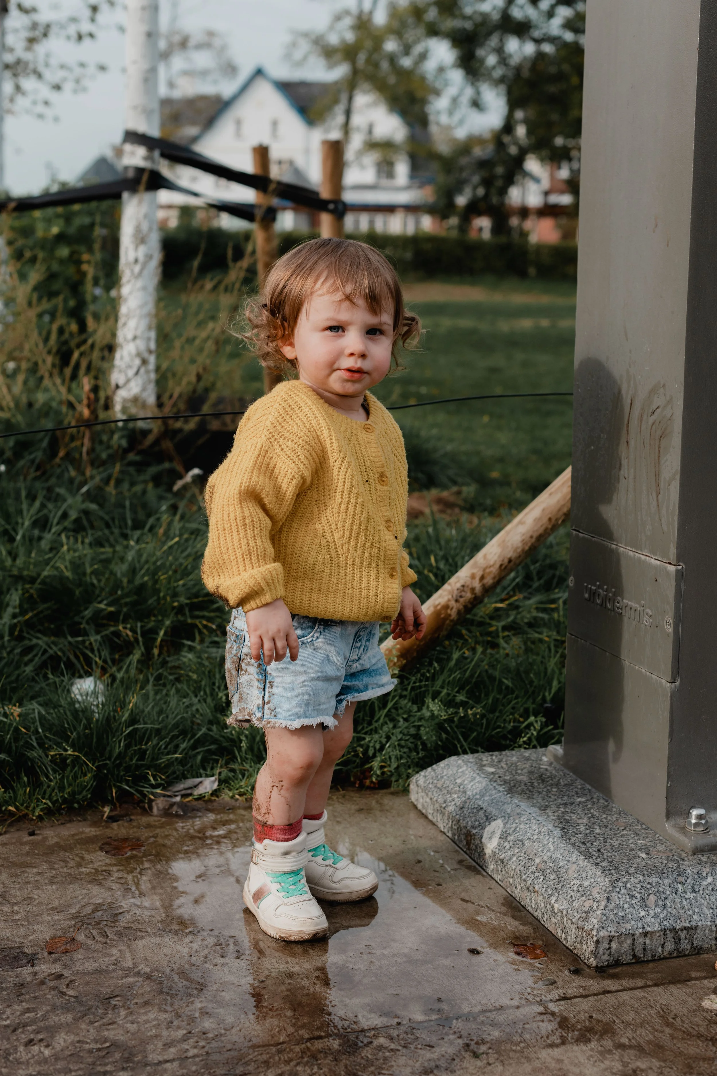 A young child standing outdoors in a muddy puddle next to a granite water fountain, wearing a yellow sweater, denim shorts, and white sneakers with teal accents, with muddy legs and socks.