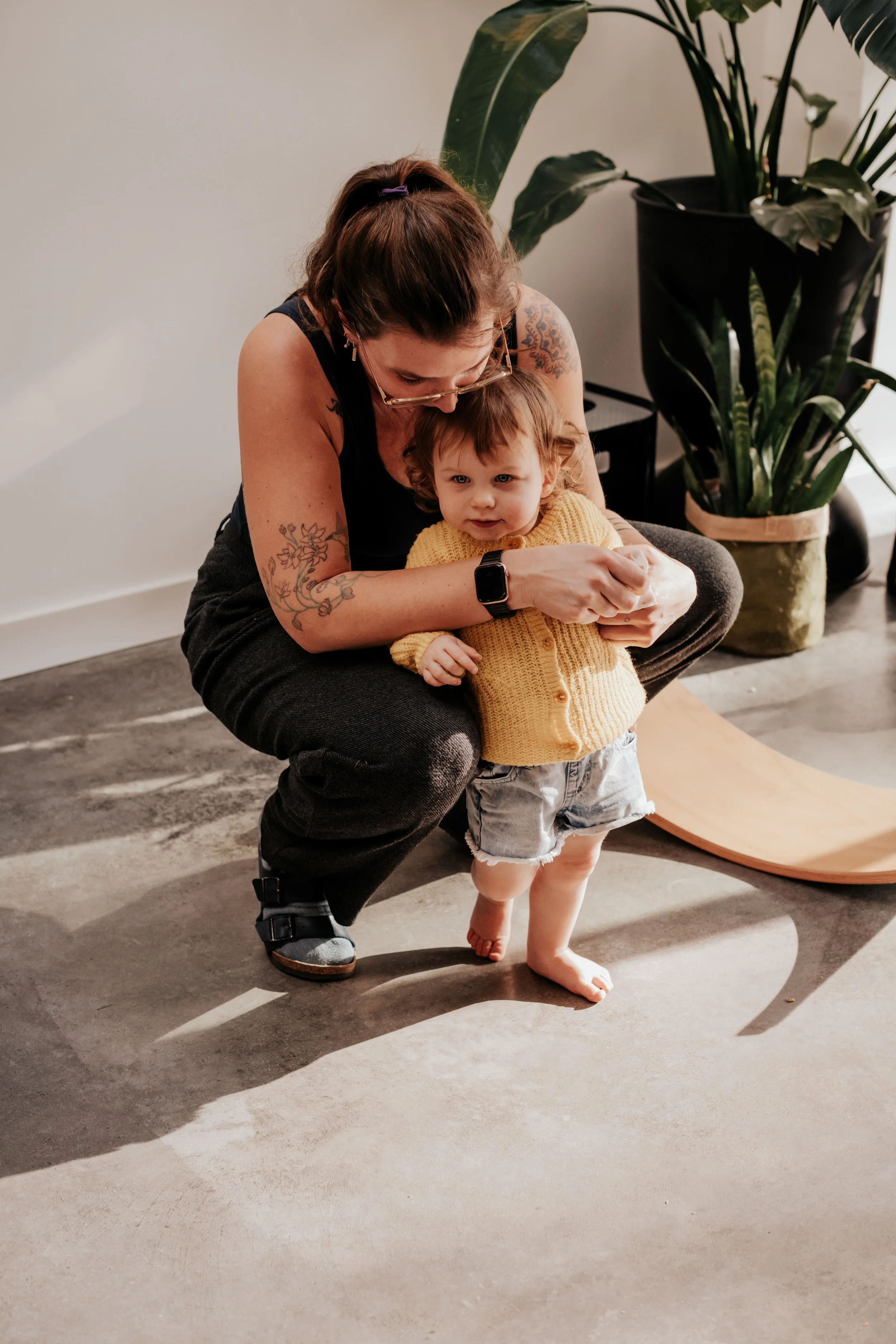 A woman with tattoos, glasses, and a purple hair tie helping a young boy with curly hair, wearing a yellow sweater, walk barefoot inside a room with a large potted plant and a modern floor lamp, casting shadows on the floor.