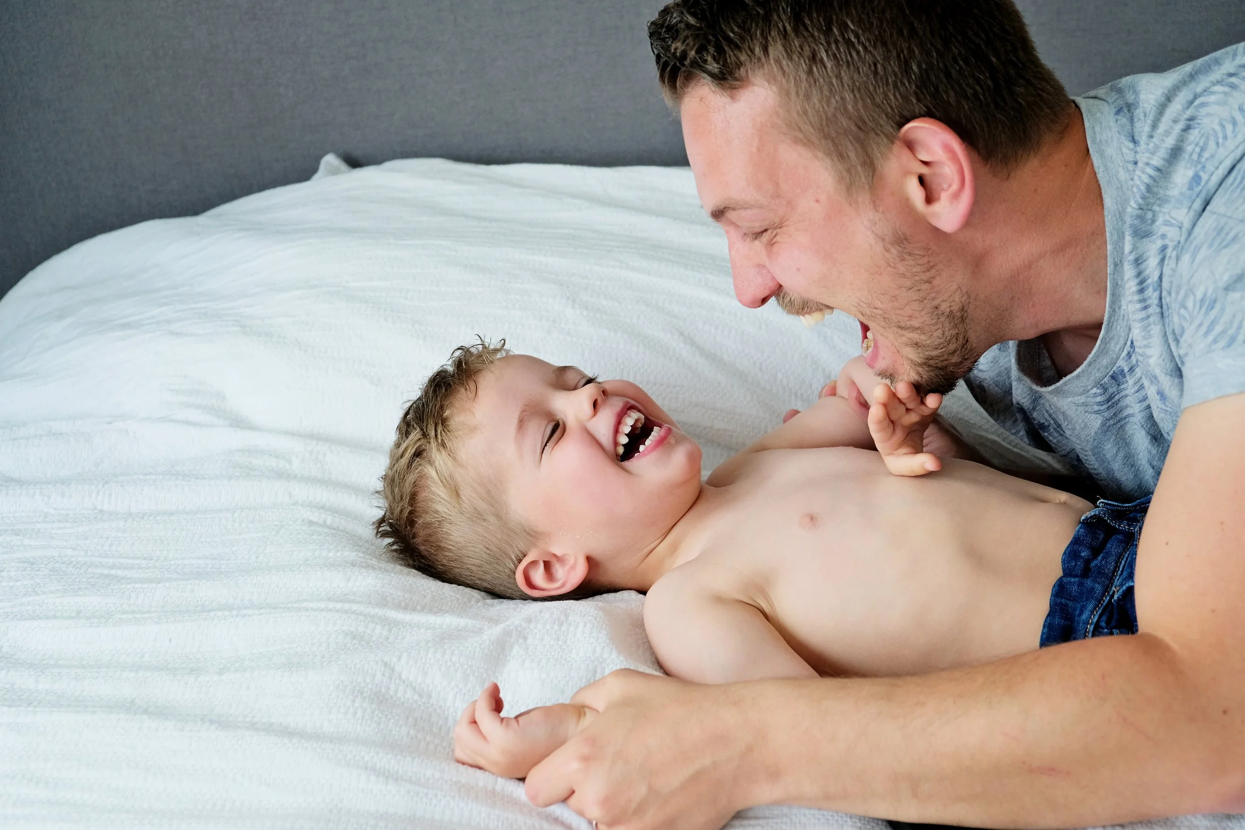A man and a young boy are playing and laughing together on a bed with a white sheet. The man is leaning over the boy, both are smiling and making playful expressions.