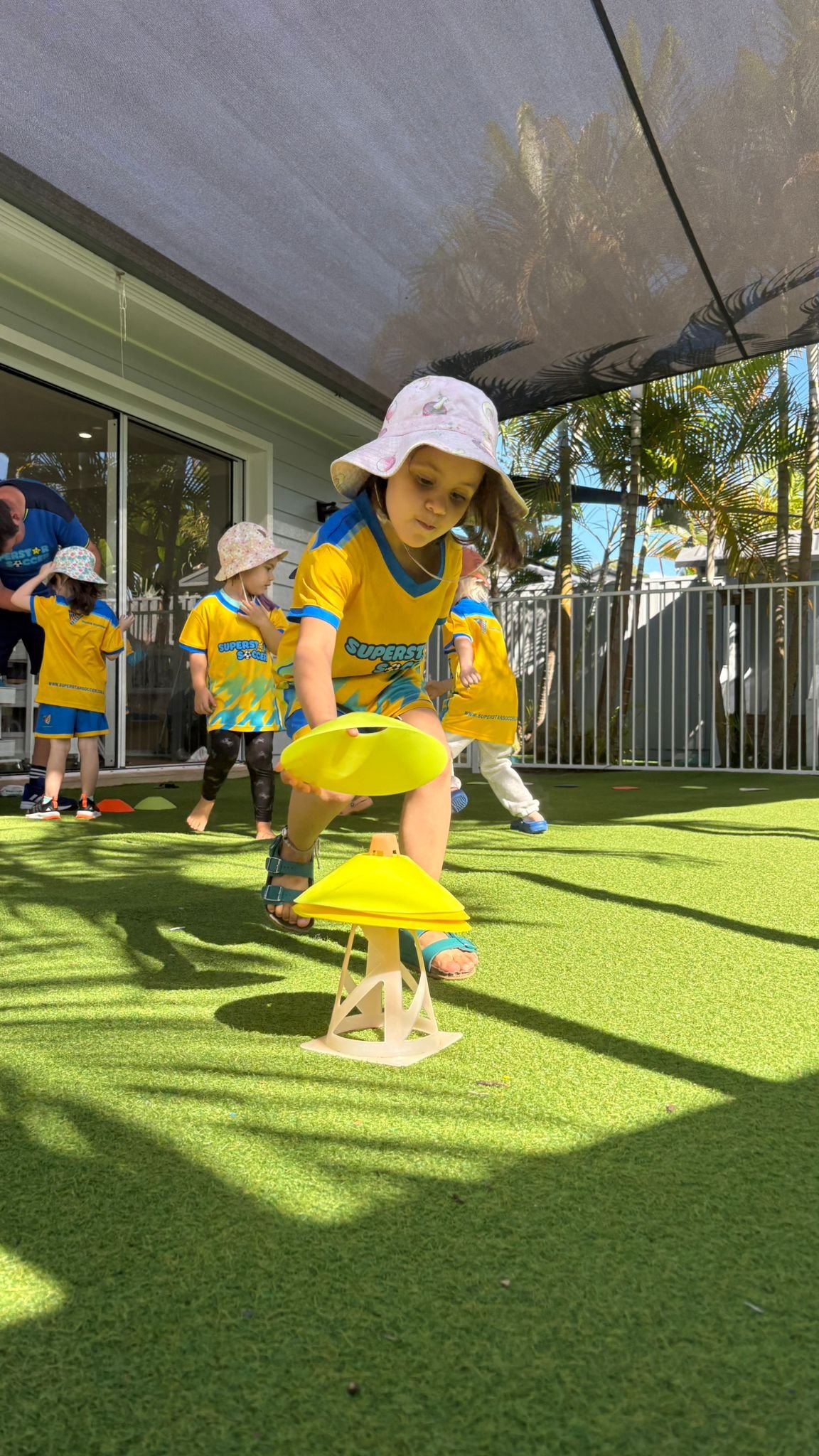 Kids participating in an outdoor game or race on a sunny day, wearing sports uniforms and hats, on a green artificial turf with a fence and trees in the background.