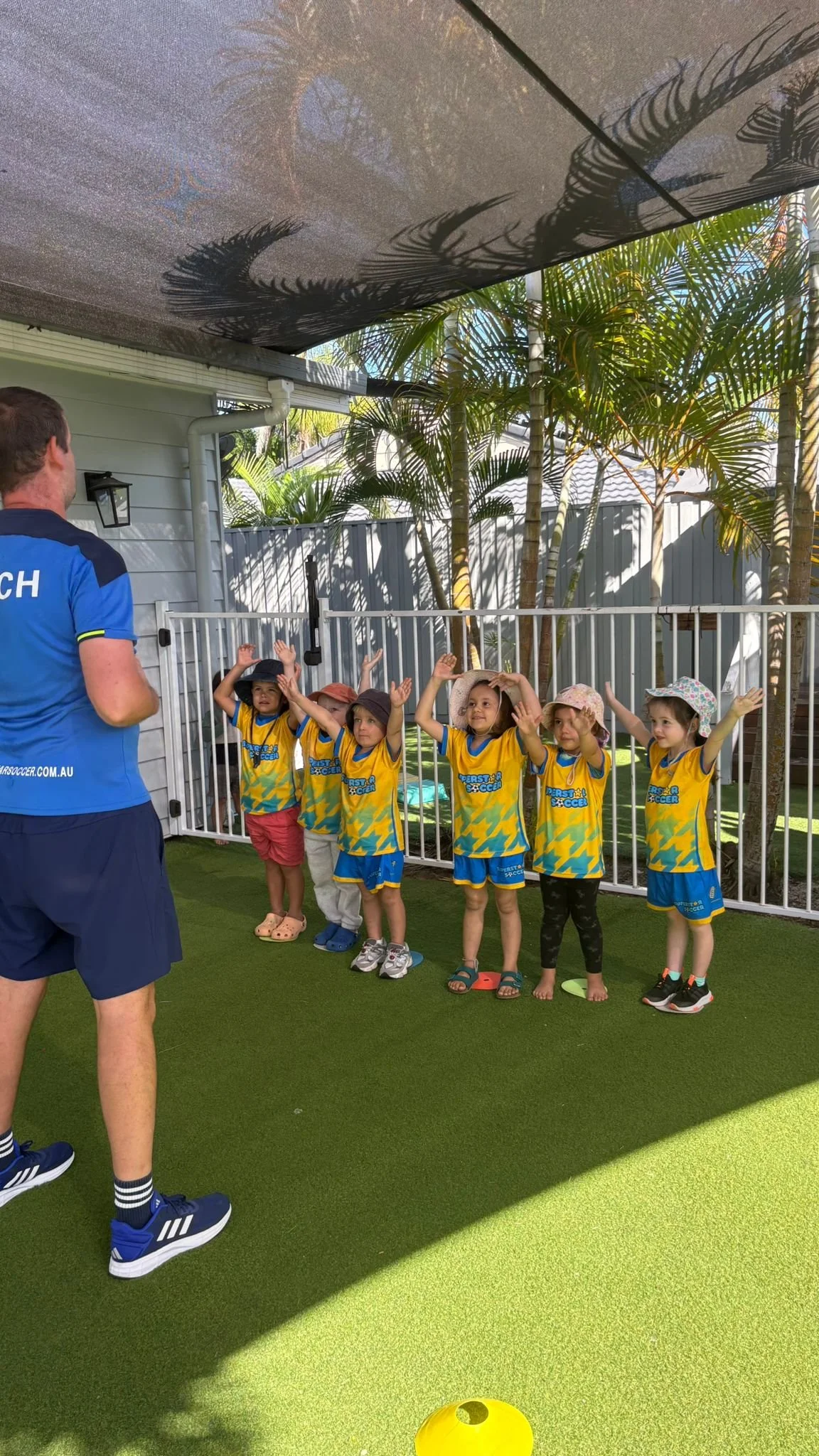 Young children in yellow and blue uniforms standing behind a fence, raising their hands during an outdoor activity, while an adult coach stands nearby under a shade with shadows cast on it from nearby plants and palm trees.