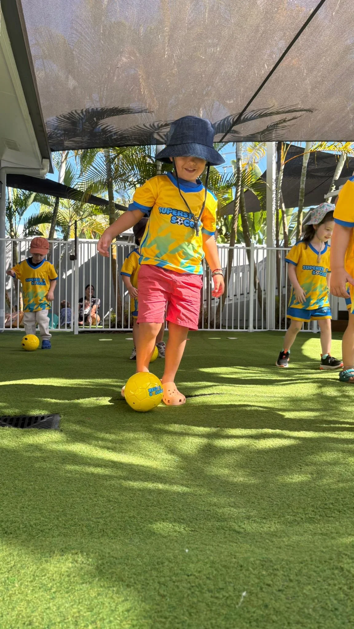 Children playing soccer in an outdoor area with artificial green turf, surrounded by a white fence and tropical plants. One child in the foreground wears a yellow shirt, pink shorts, a wide-brimmed hat, and Crocs, controlling a yellow soccer ball. Other children are also wearing yellow shirts, some with hats, participating in the activity.
