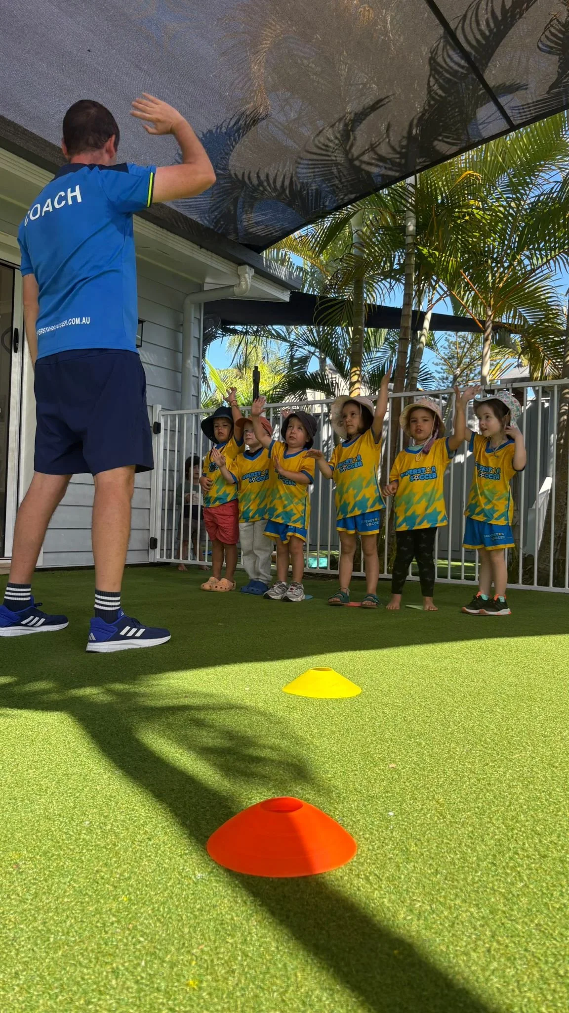 A coach leading a group of young children in a soccer training session on an outdoor artificial turf pitch, with orange cones on the ground and palm trees in the background.