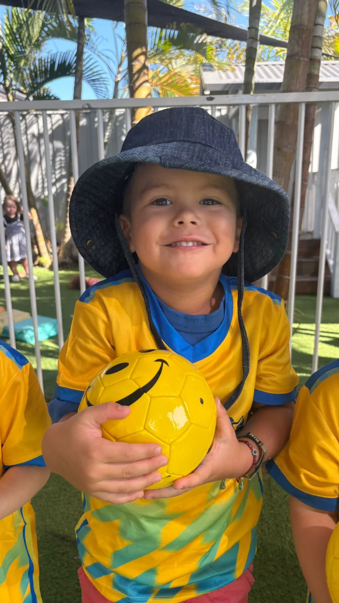A young boy smiling while holding a yellow soccer ball with a smiley face design, wearing a yellow sports jersey and a wide-brimmed hat, outdoors in a sunny setting.
